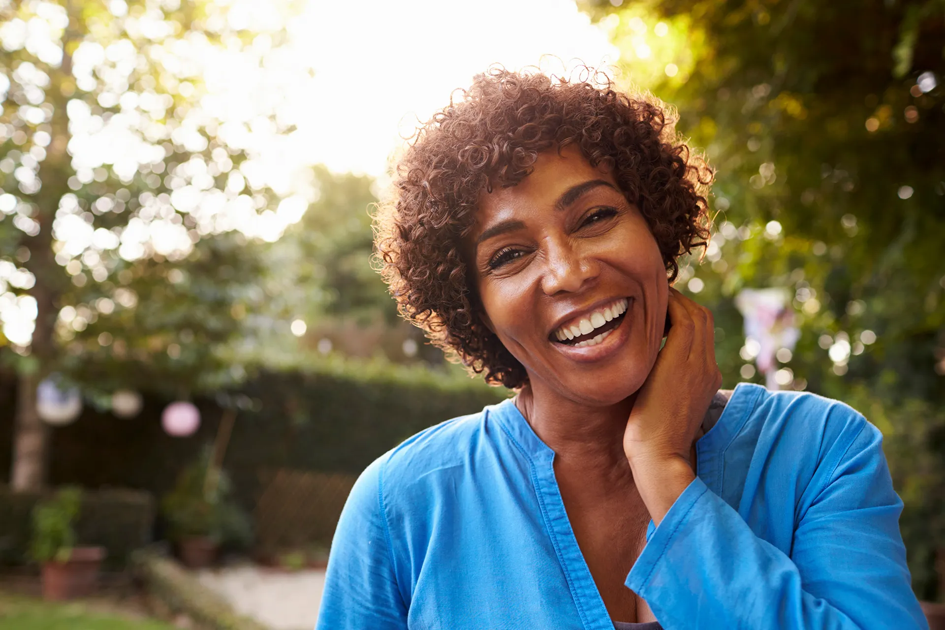 Smiling woman in blue shirt enjoying a sunny day outdoors in a garden.