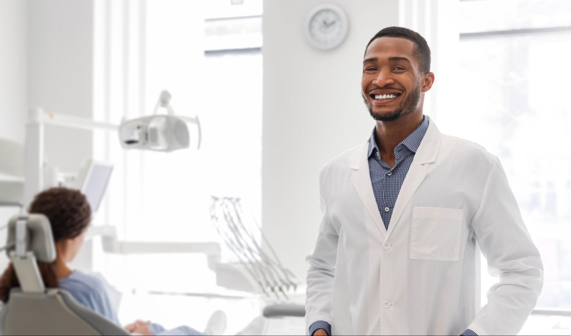 Smiling dentist with a female patient in the background sitting in an exam chair. 