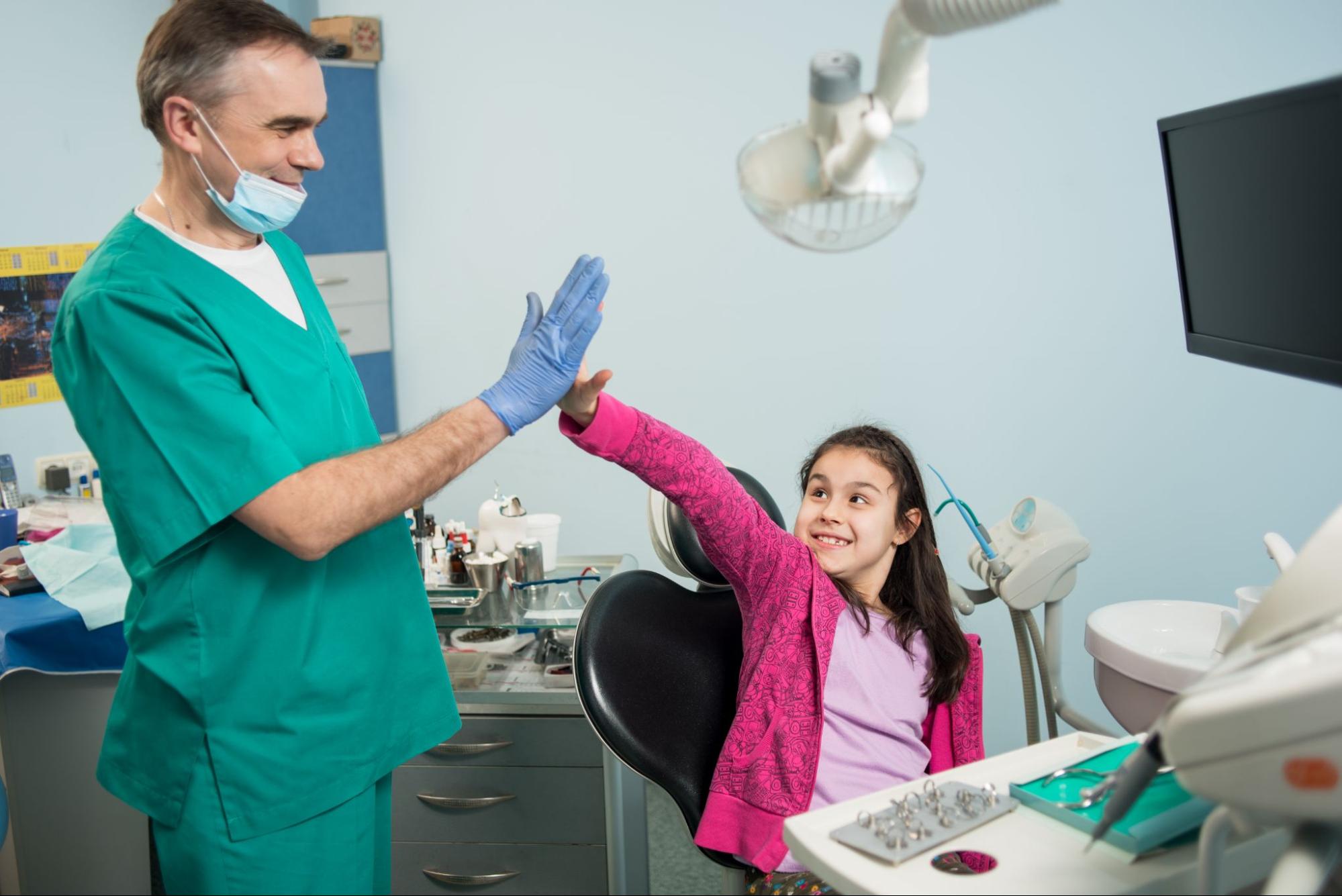 A young girl high-fives her senior pediatric dentist after a successful first dental visit, celebrating a positive experience during her checkup.