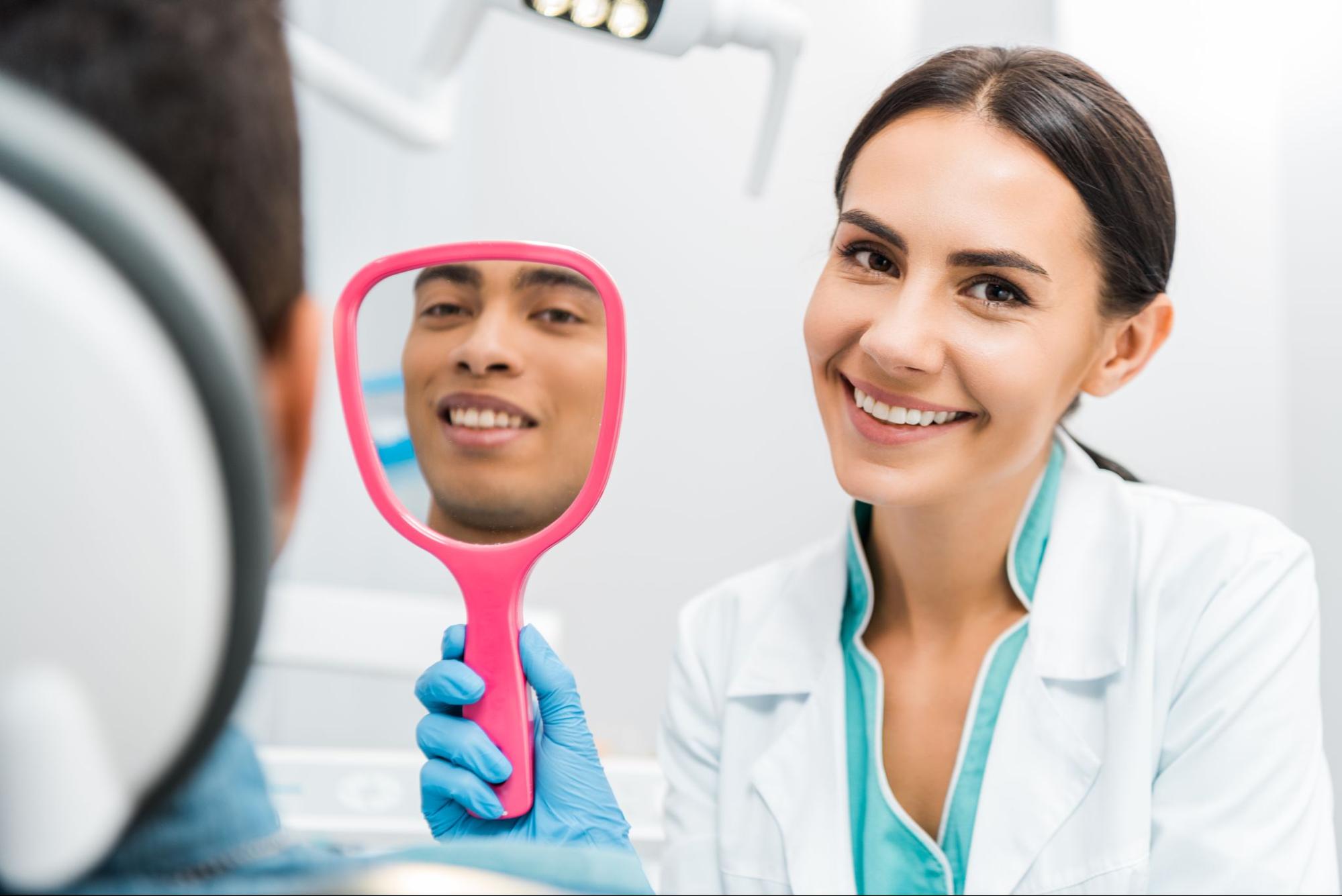 Man smiling in a handheld mirror next to a female dentist. 