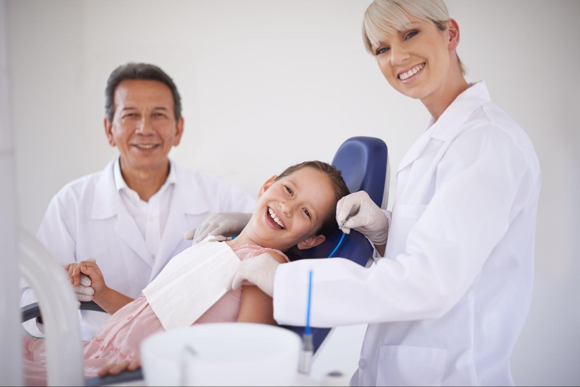 Young girl in a dental exam chair smiling next to a male and female dentist. 