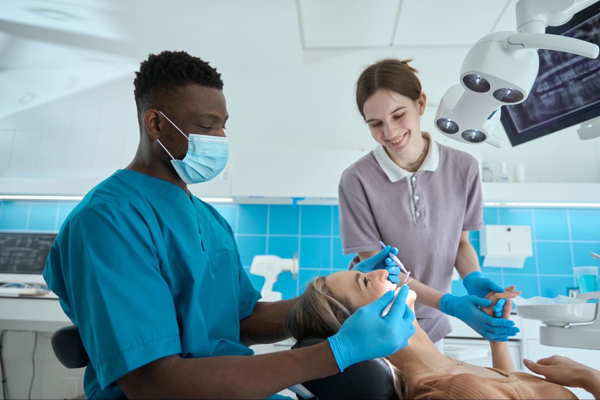 In a modern dental clinic, a Black male endodontist examines a woman's teeth while his female assistant comforts the patient by holding her hand.