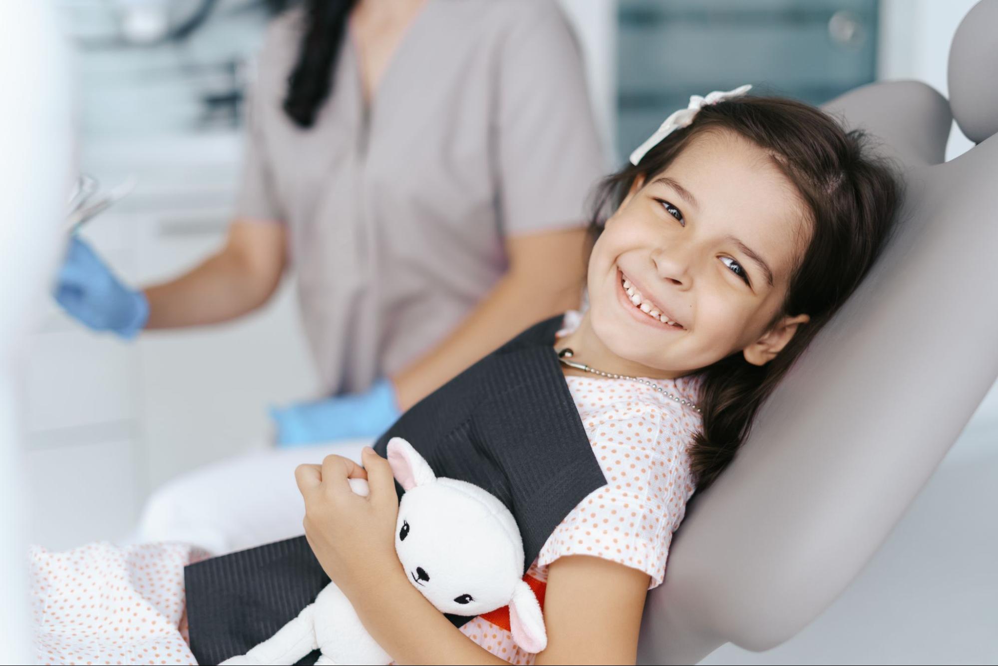 Young girl in an exam chair holding a stuffed animal and smiling. 