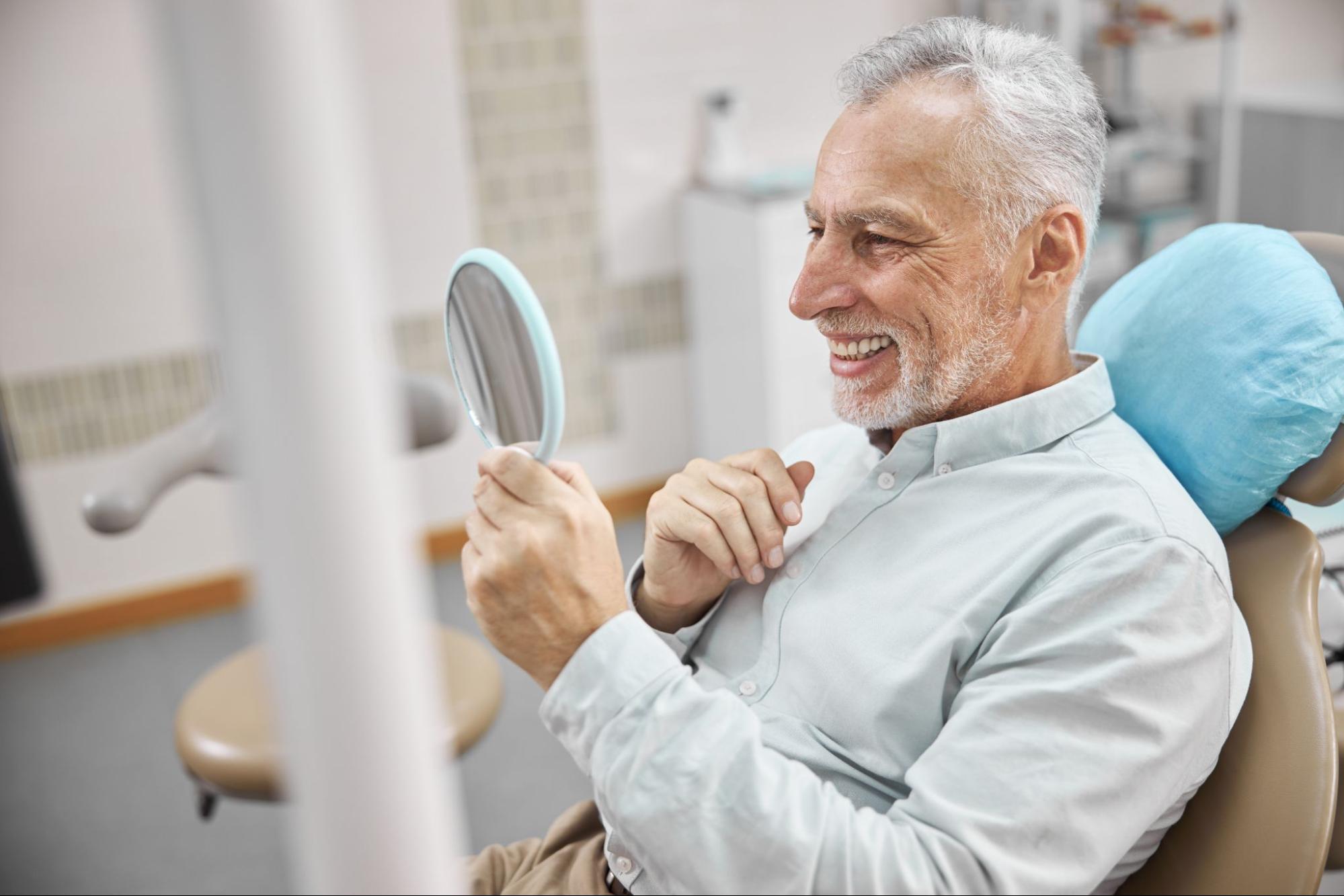 A happy older man smiles as he examines his teeth in the mirror, satisfied with his partial dental implants.