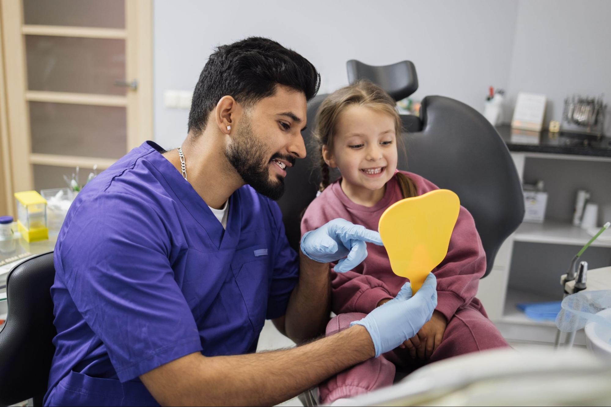 A cheerful young girl smiles at her reflection in a mirror, delighted by her bright, healthy teeth, while her male dentist shows her the results.