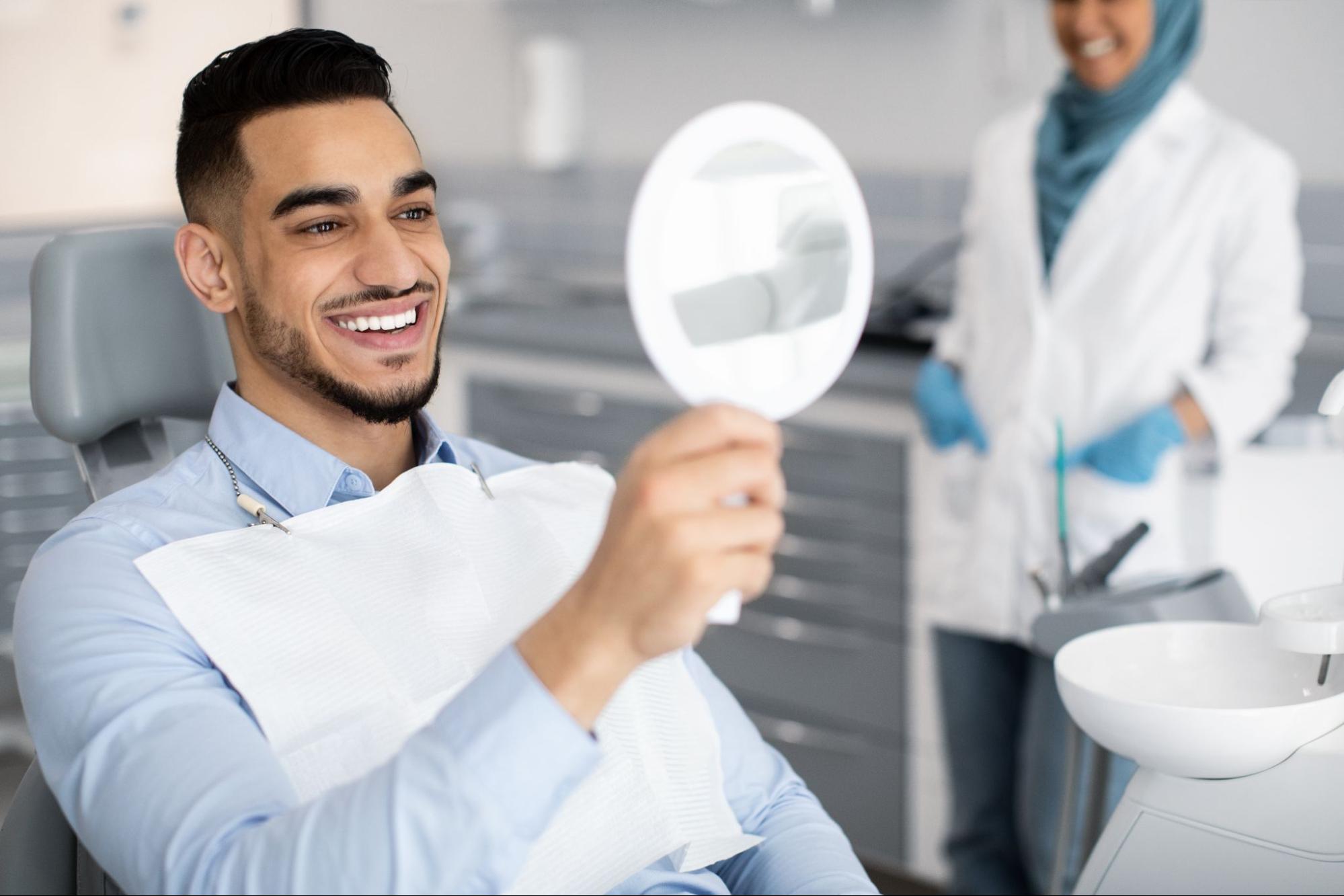 Man with a white smile sitting in an exam chair holding up a mirror next to a female dentist. 