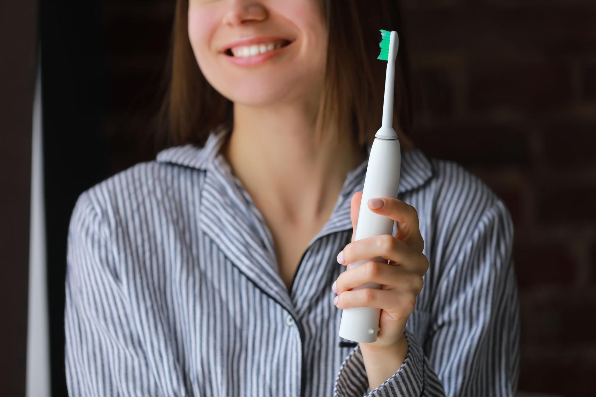 A cropped shot of a smiling woman in blue and white striped pajamas holding an ultrasonic electric toothbrush.