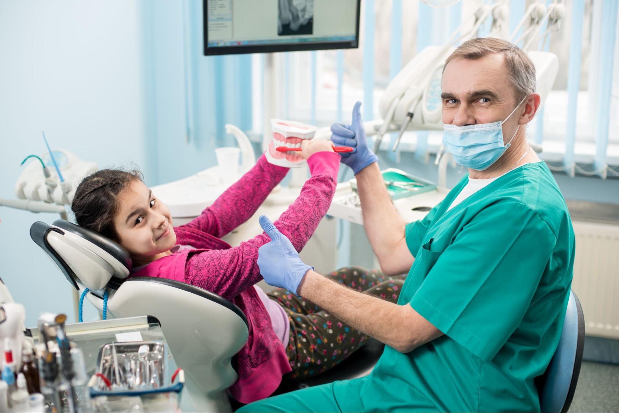 Dentist wearing a mask with both thumbs up next to a young girl brushing a model of a mouth. 