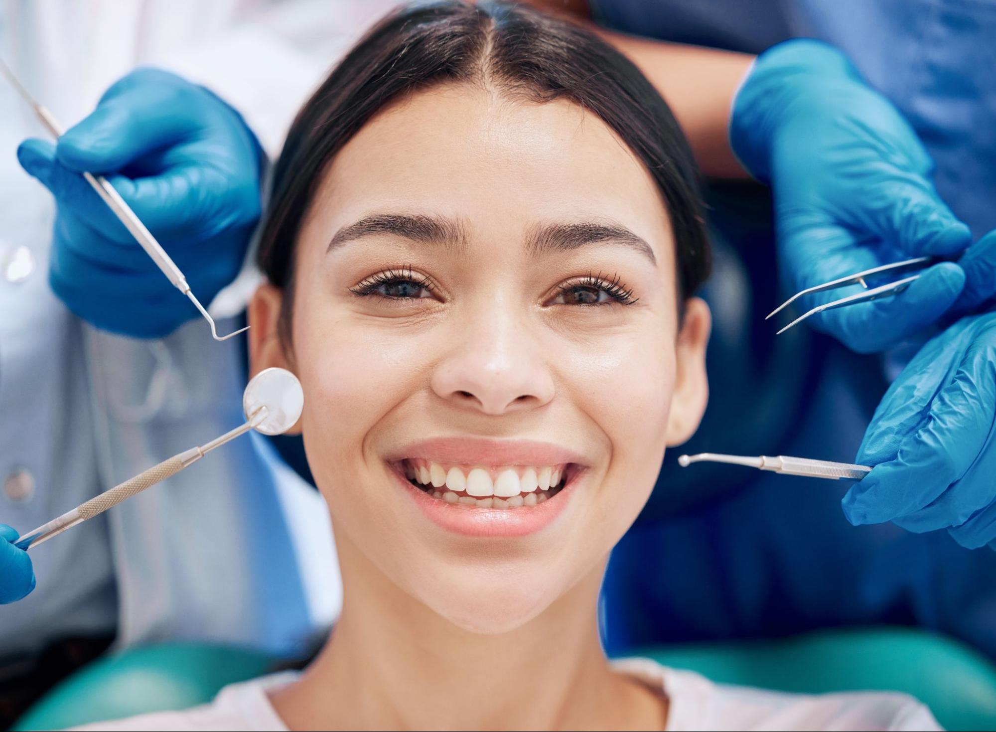 A close-up of a joyful woman smiling after a dental checkup, with hands in blue gloves holding dental tools visible around her.
