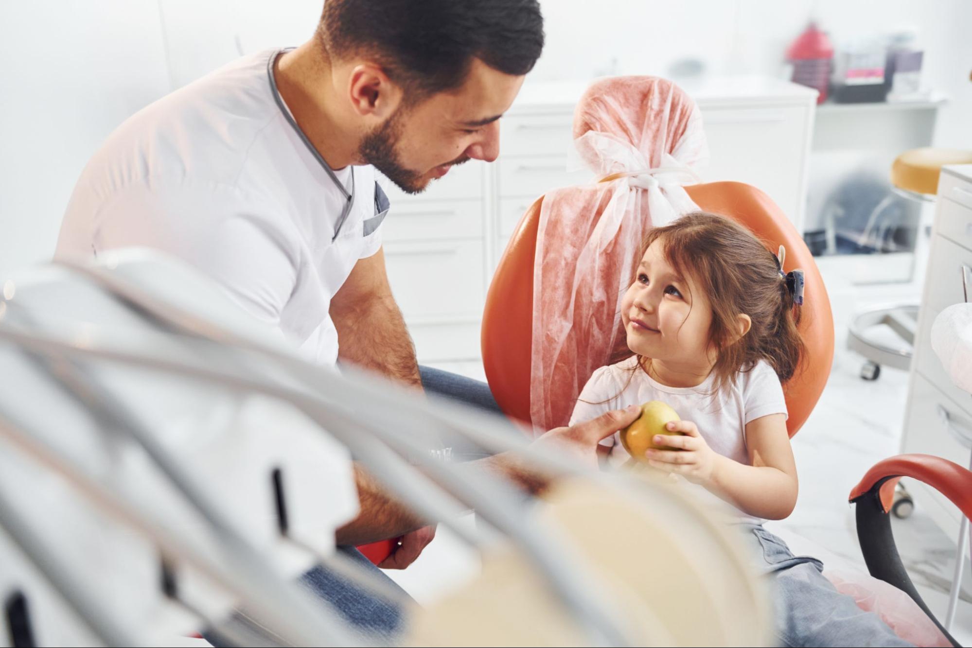 A young girl sits in the dental chair during a clinic visit, and the dentist gives her an apple as he prepares her for a checkup.
