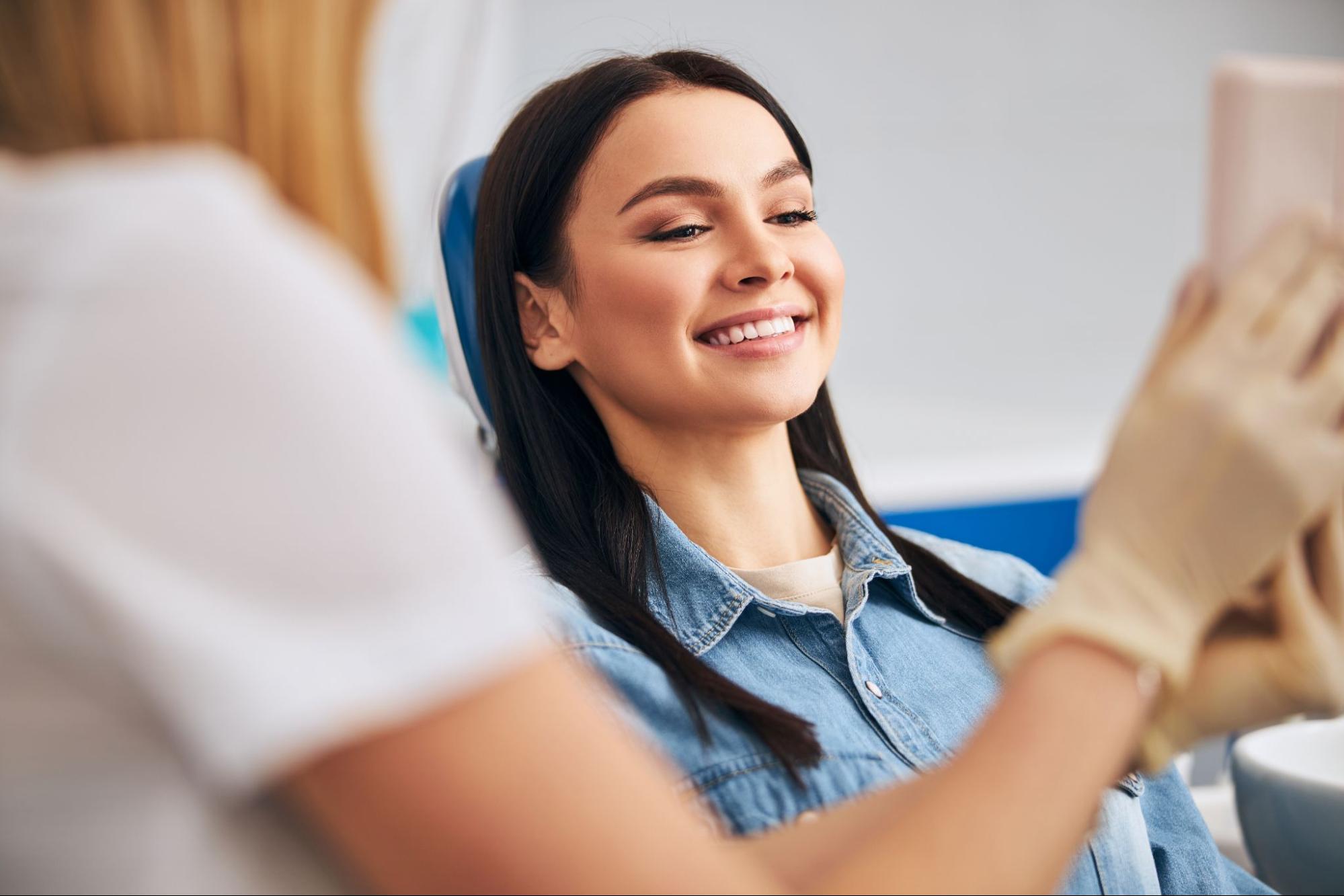 Woman smiling and looking at a mirror while in an exam chair next to a female dentist. 