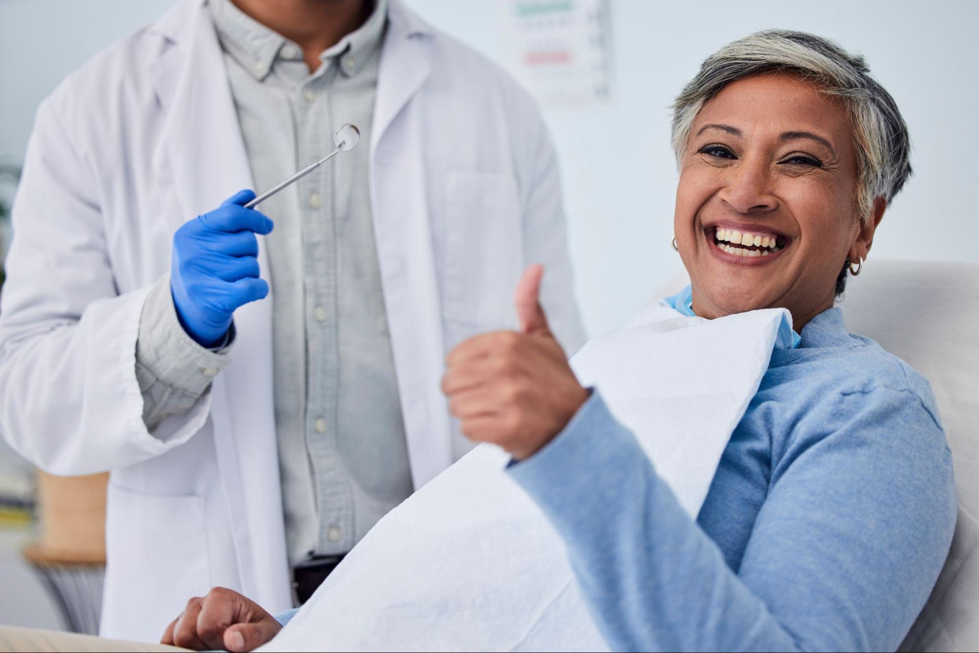 Woman smiling and giving a thumbs up while sitting next to a dentist.