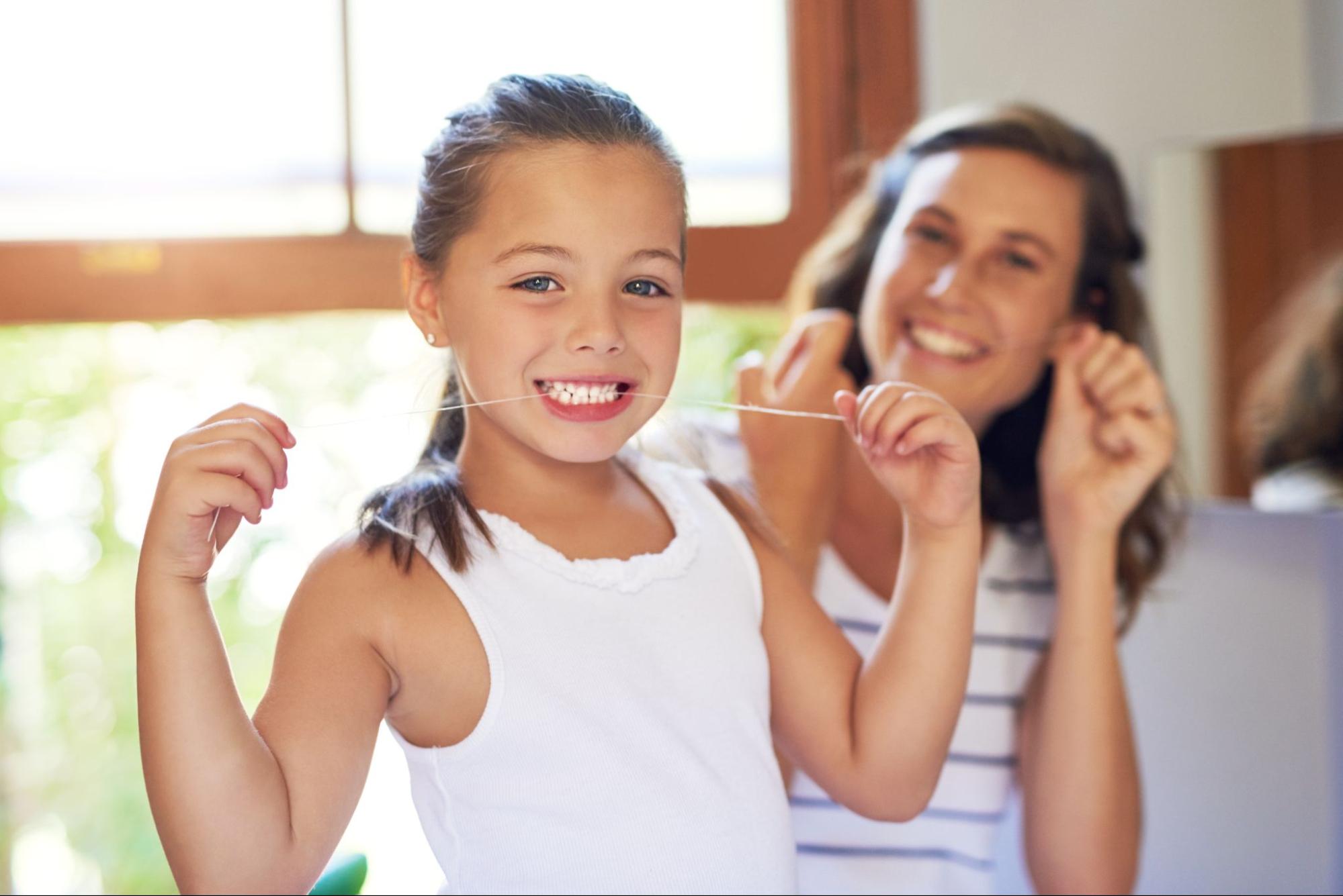 A happy young girl flosses her teeth with a smile as her mother proudly looks on from behind.