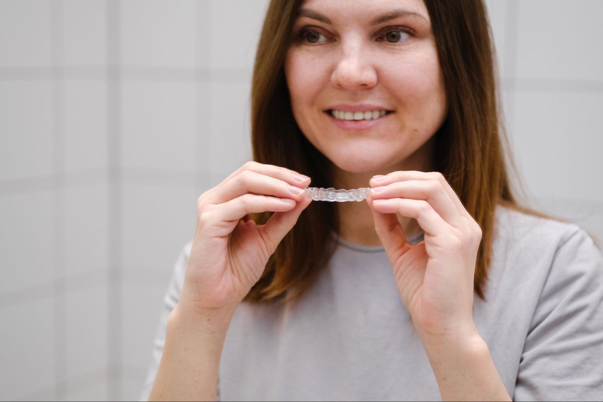 A woman stands in front of a mirror holding clear plastic aligners, preparing to put them on her teeth.