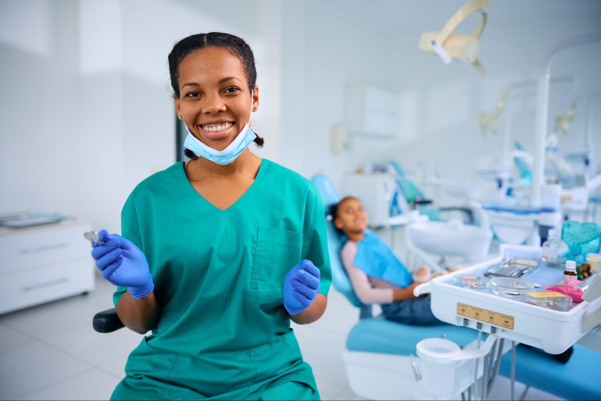 A happy female dentist smiles at the camera, with a young patient in the background, ready for treatment.