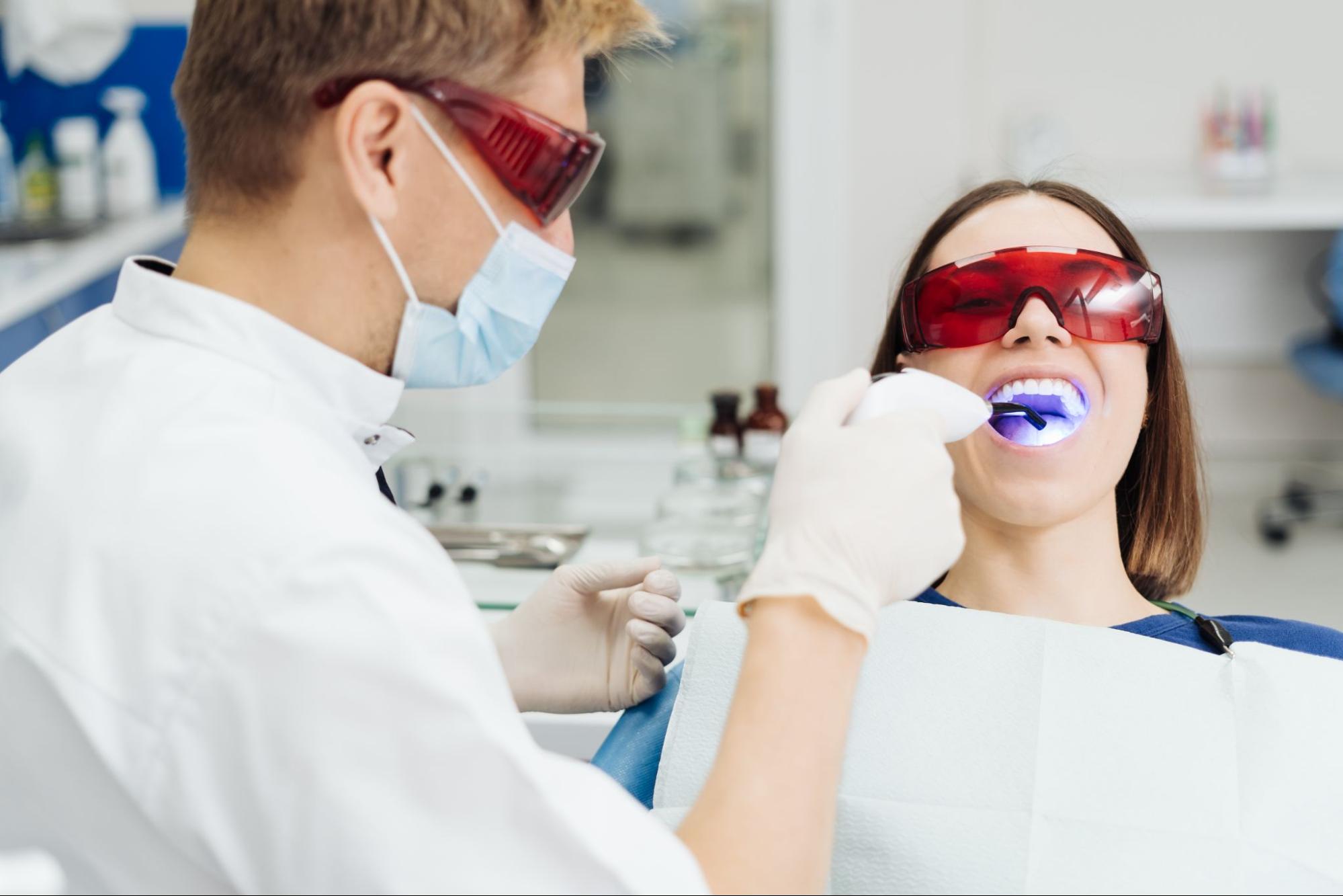 A young woman undergoes dental treatment from a male dentist. Both wear protective red glasses to shield their eyes from the curing light used for dental procedures.