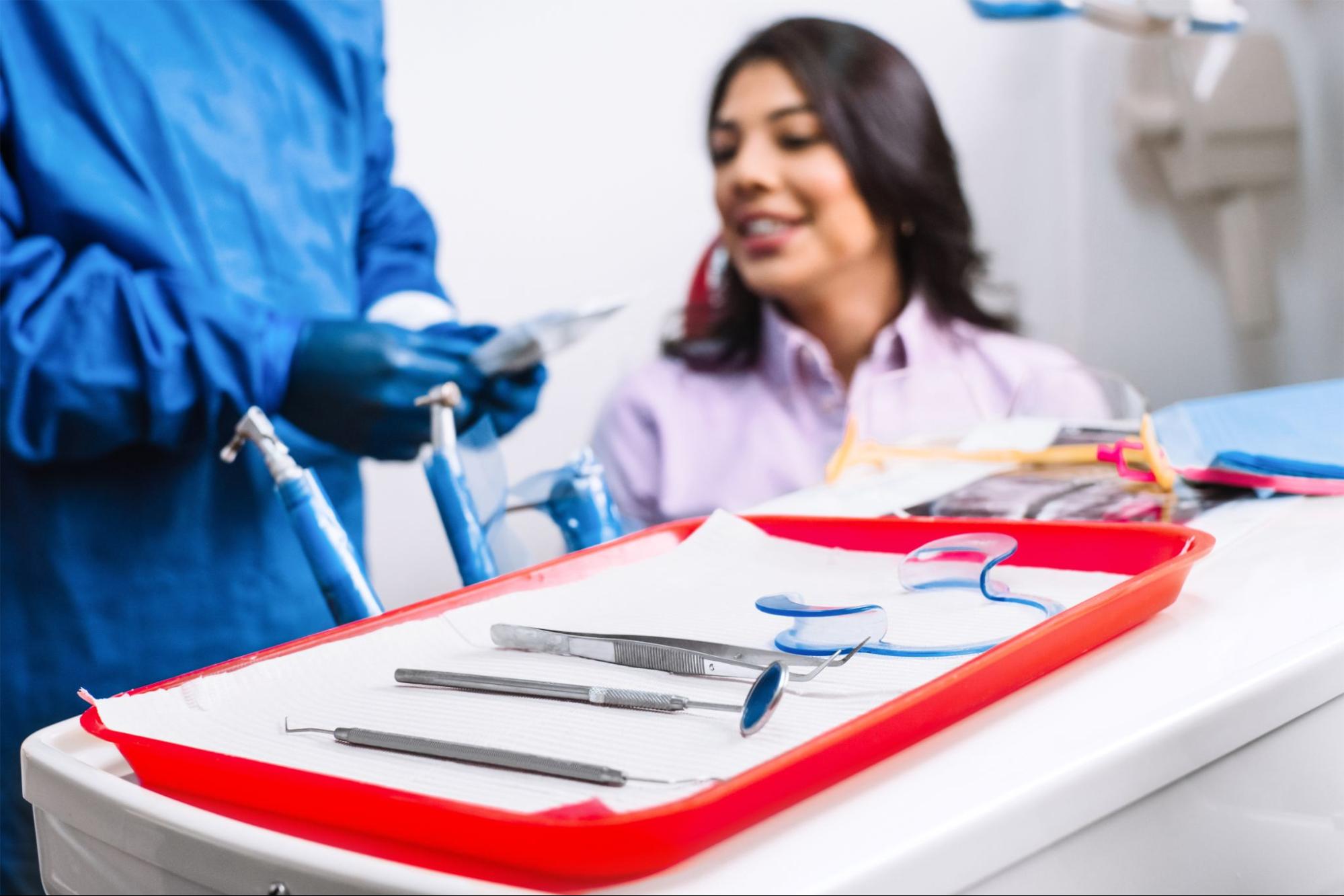 Dental instruments and tools are laid out before a smiling female patient while an unidentified dentist in blue PPE holds a small sachet containing dental materials.