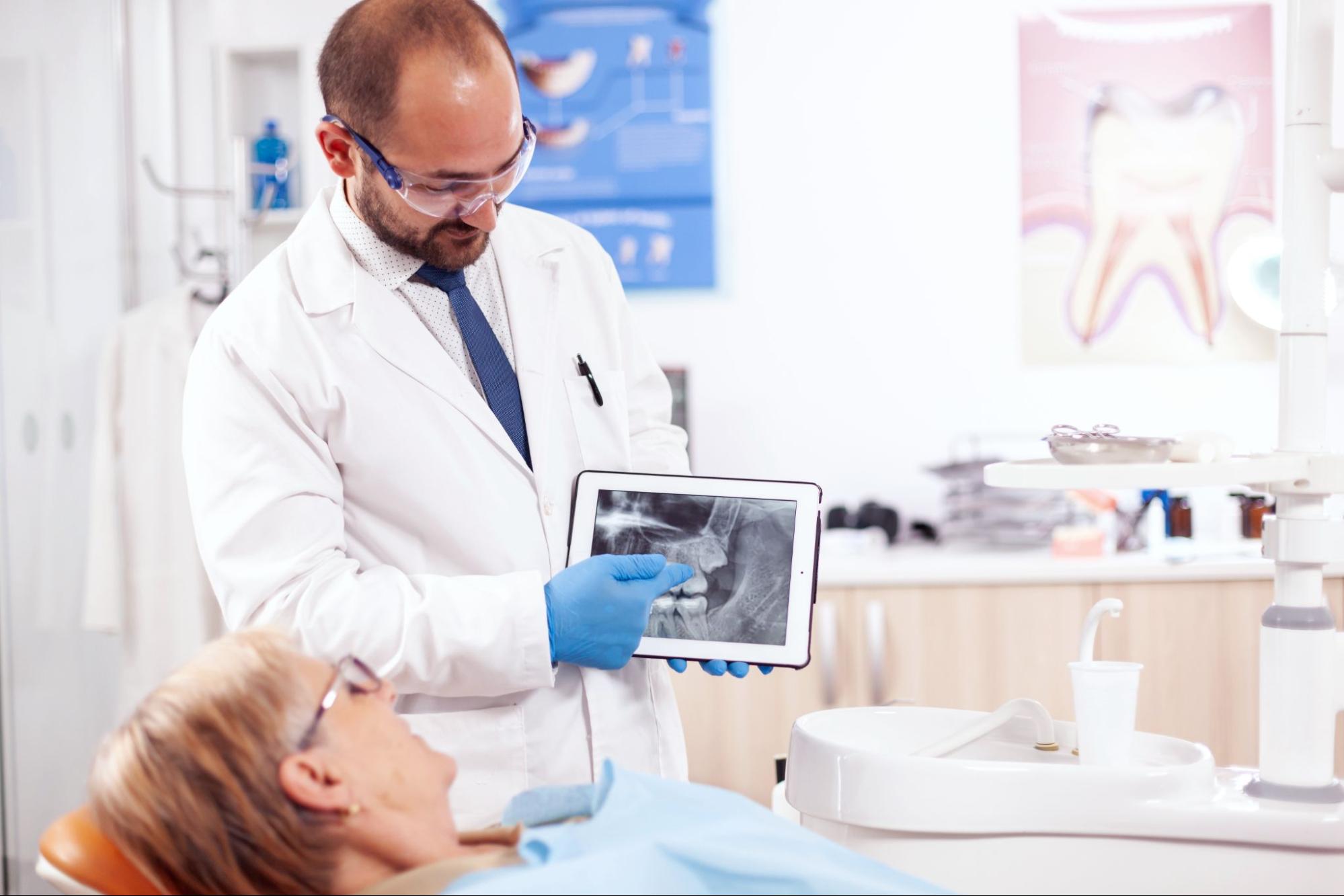 A male bearded dentist wearing glasses shows a patient's dental X-ray on a tablet while explaining the diagnosis in a clean clinic; posters of teeth and dental equipment are visible in the background.