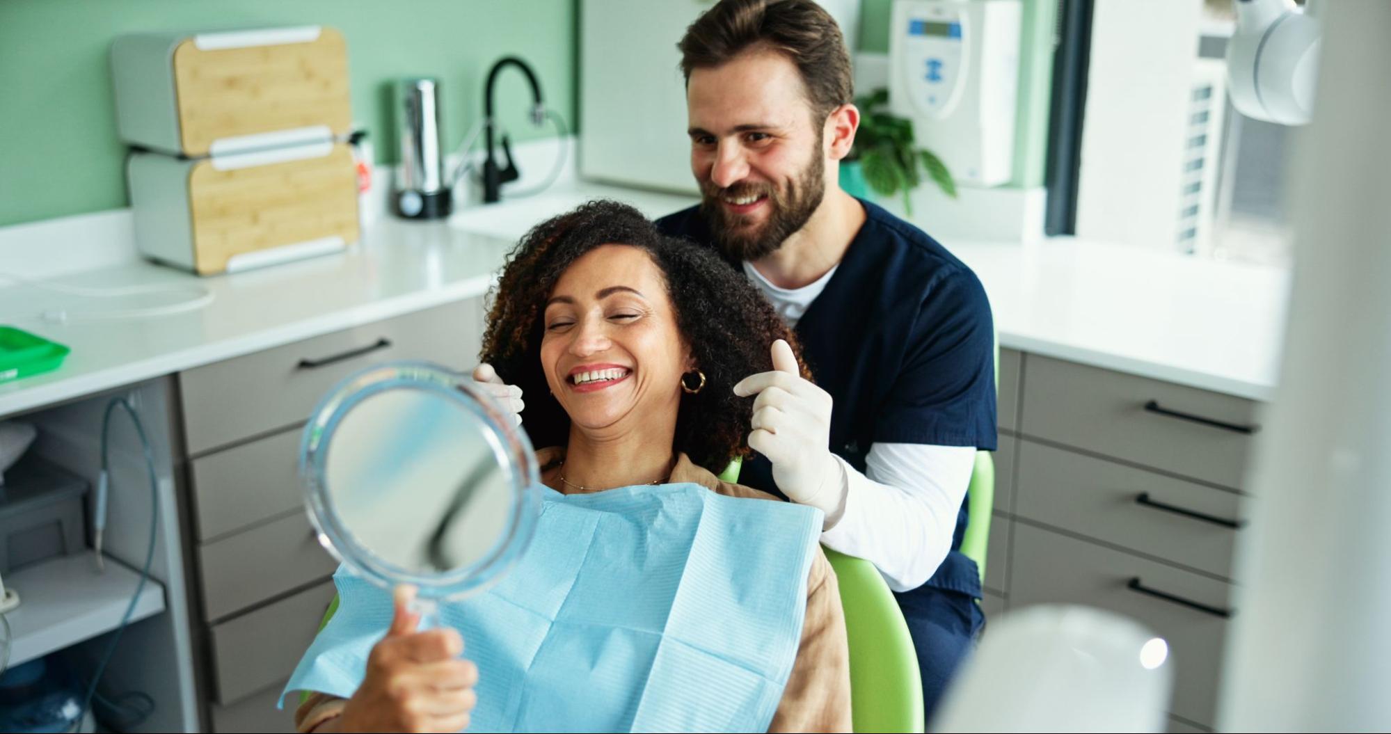 Woman looking at her teeth in a mirror next to a dentist. 