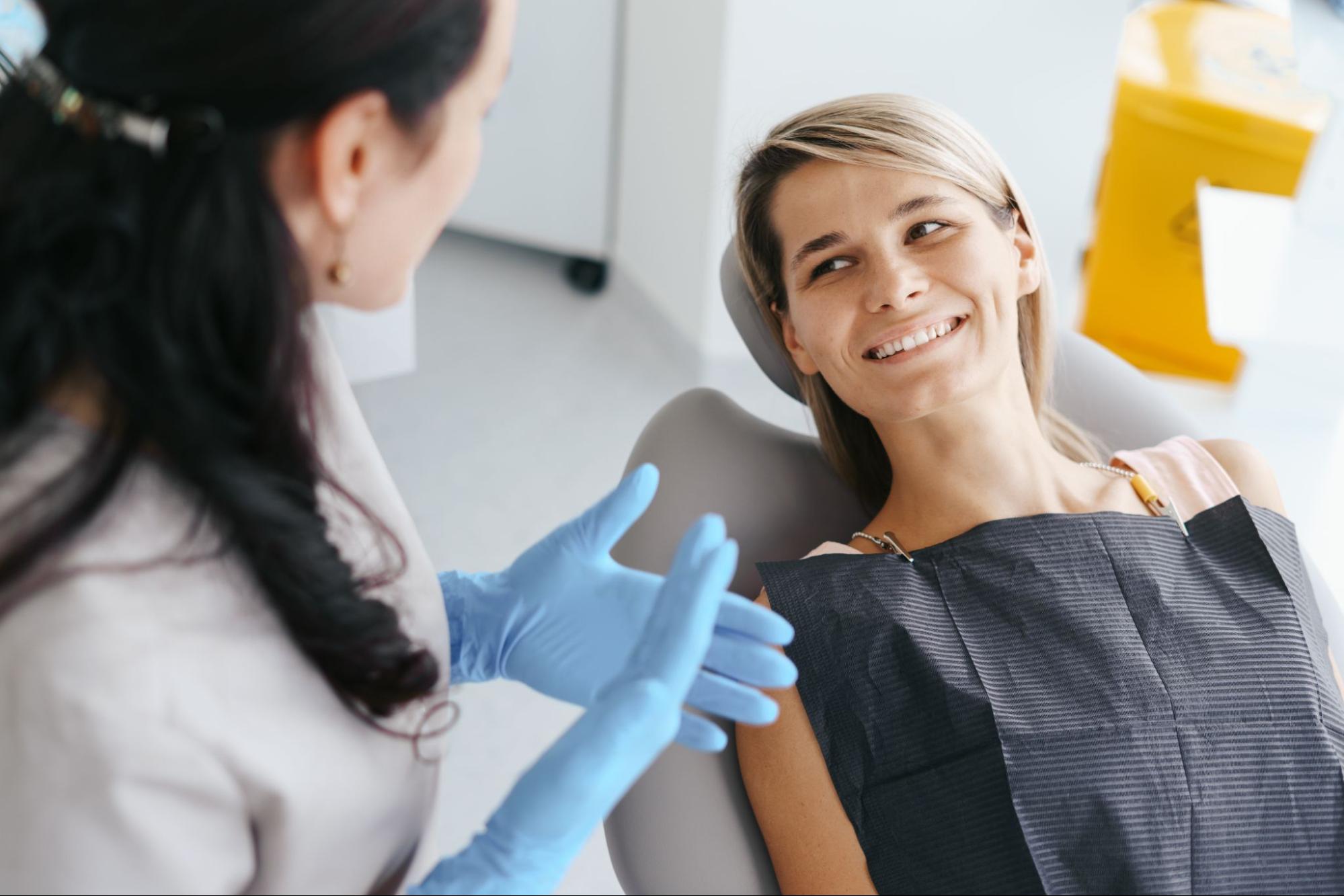 A dentist wearing blue gloves talks to a smiling patient seated in a dental chair.