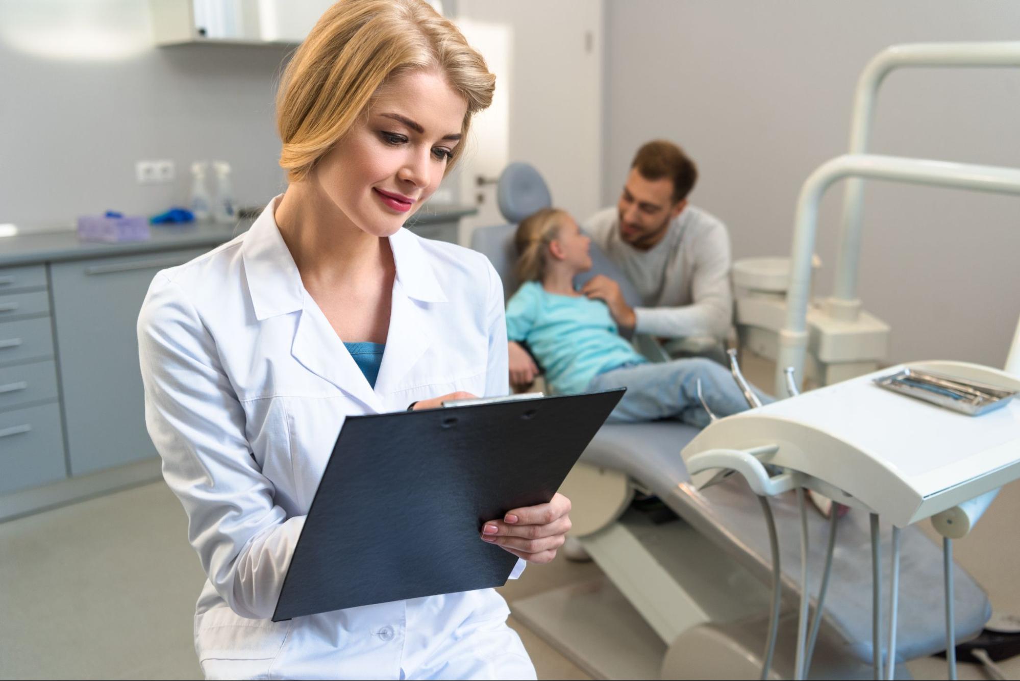 A smiling female dentist writes the next dental checkup appointment on a clipboard while a young child and her father sit in the background.