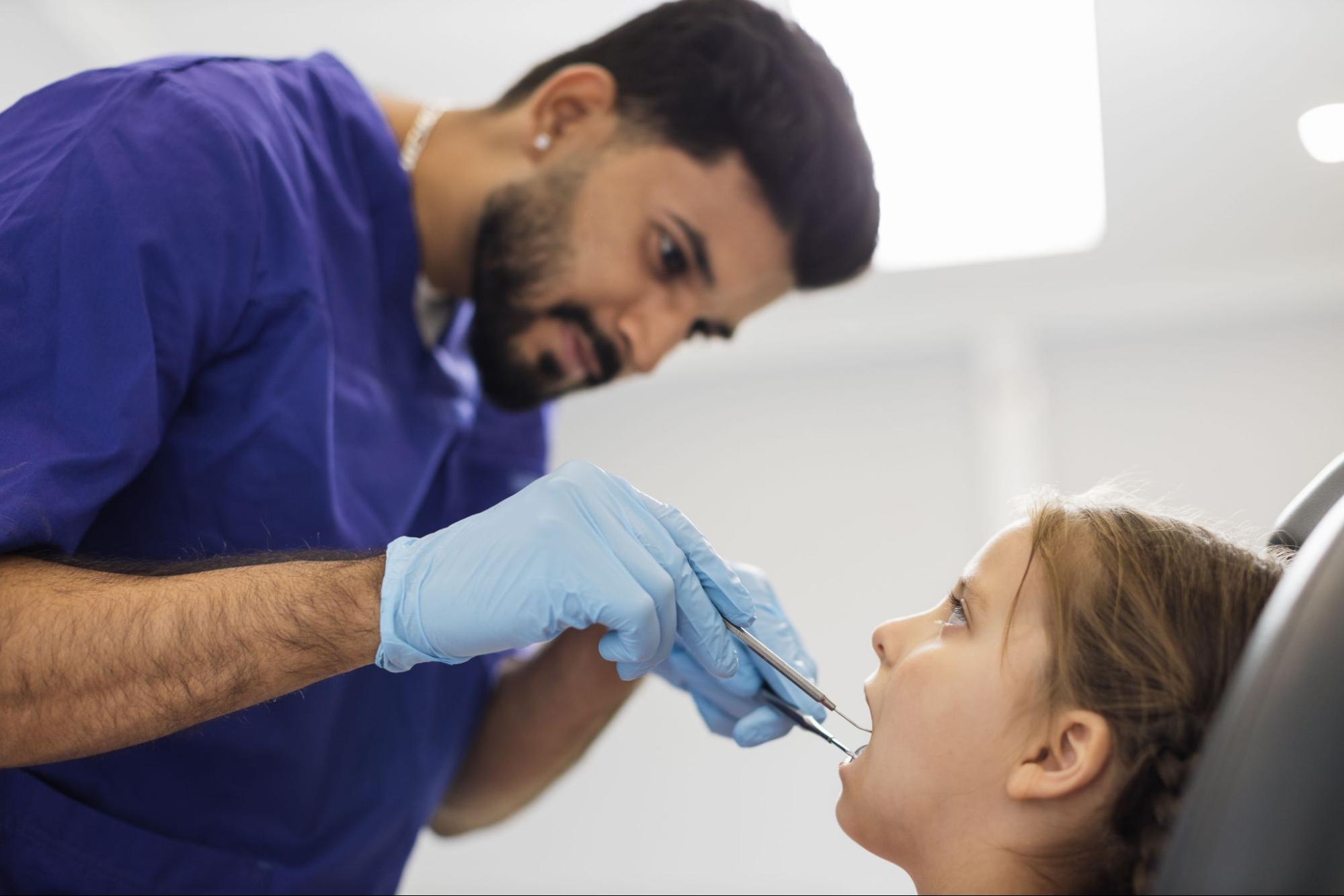 A male Hindi dentist in uniform and gloves examines a child patient's cavity during a dental procedure.
