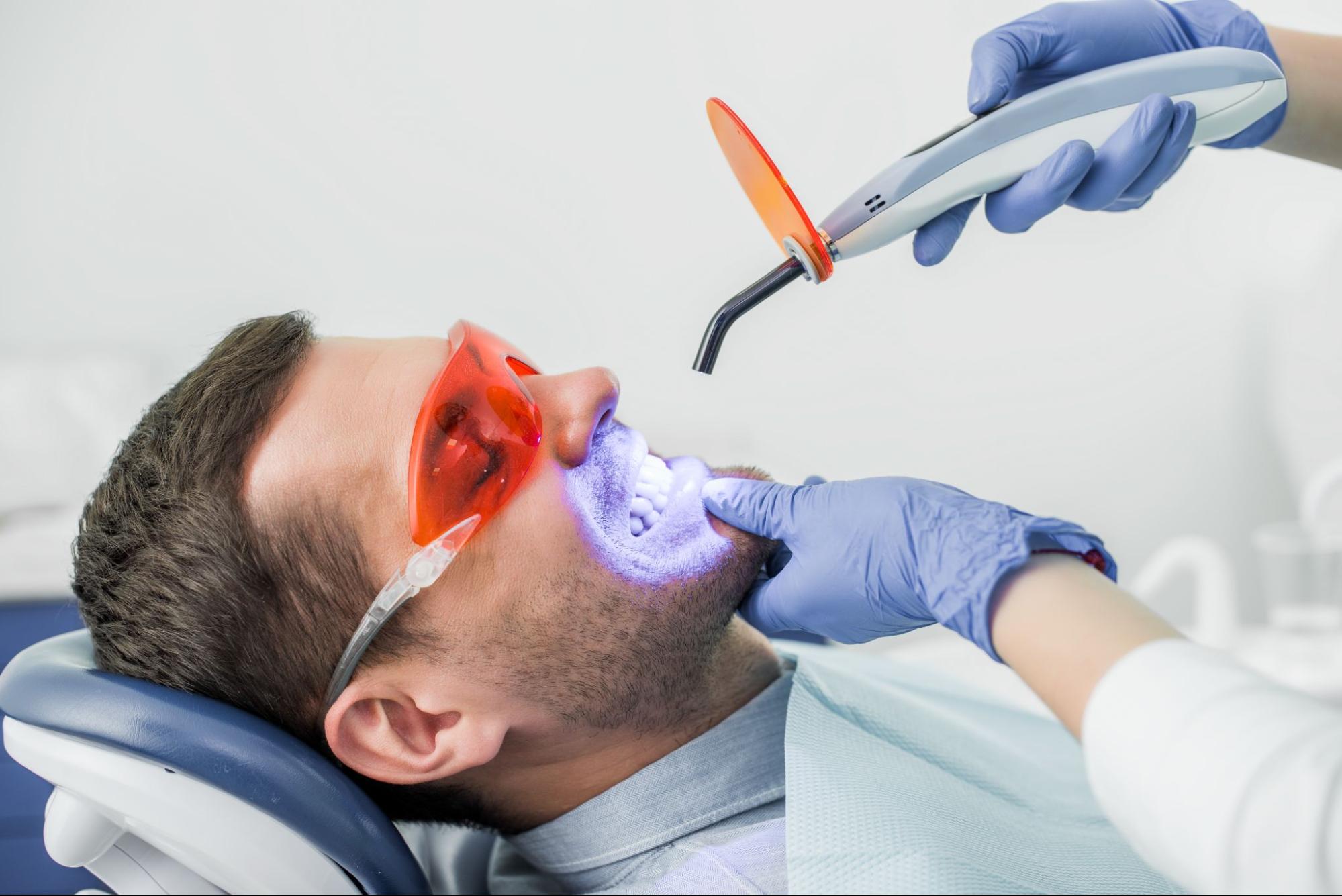 A cropped view of a dentist performing a teeth bleaching procedure on a male patient wearing protective glasses.