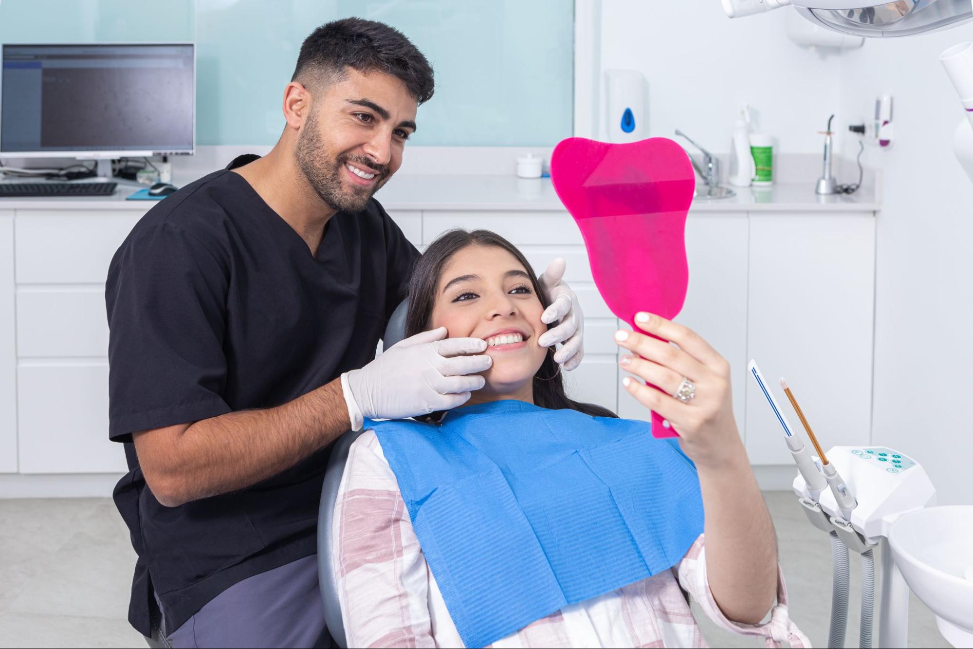 A dentist gently holds a female patient’s cheeks as she admires her newly bonded teeth in a pink hand mirror, smiling with satisfaction