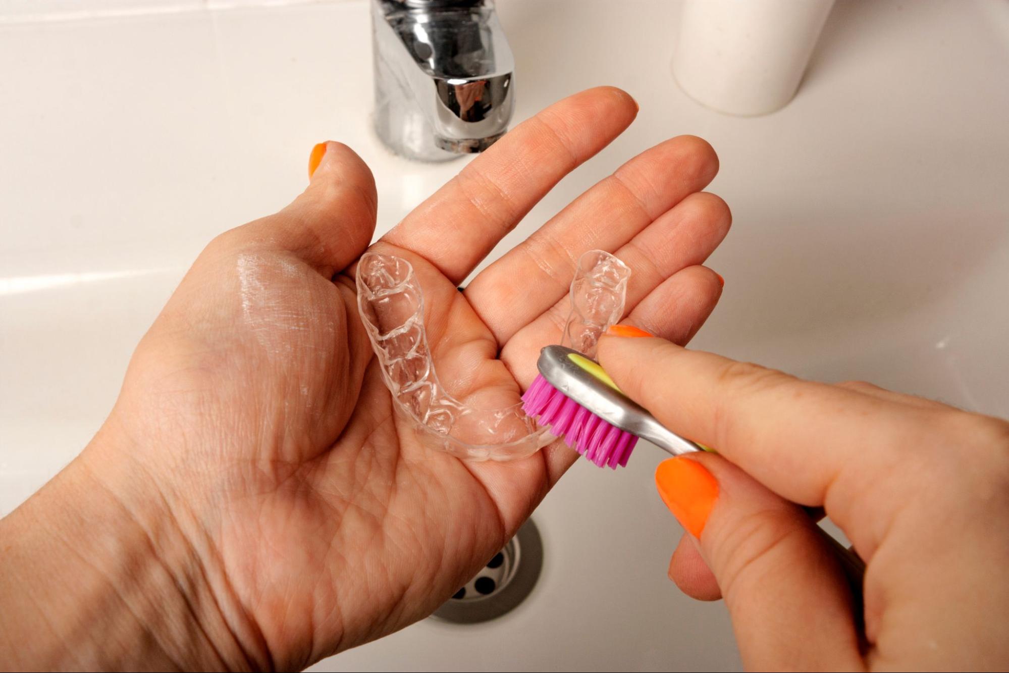 A man holds an Invisalign® aligner, cleaning it with a soft toothbrush over a sink.