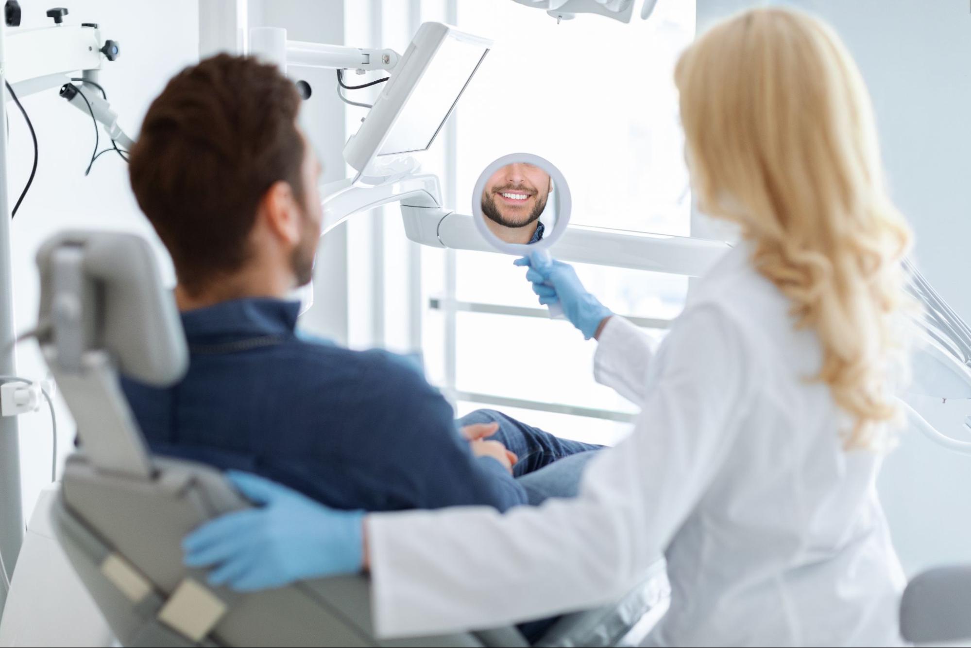 Man smiling in a mirror next to a female dentist while sitting in an exam chair.