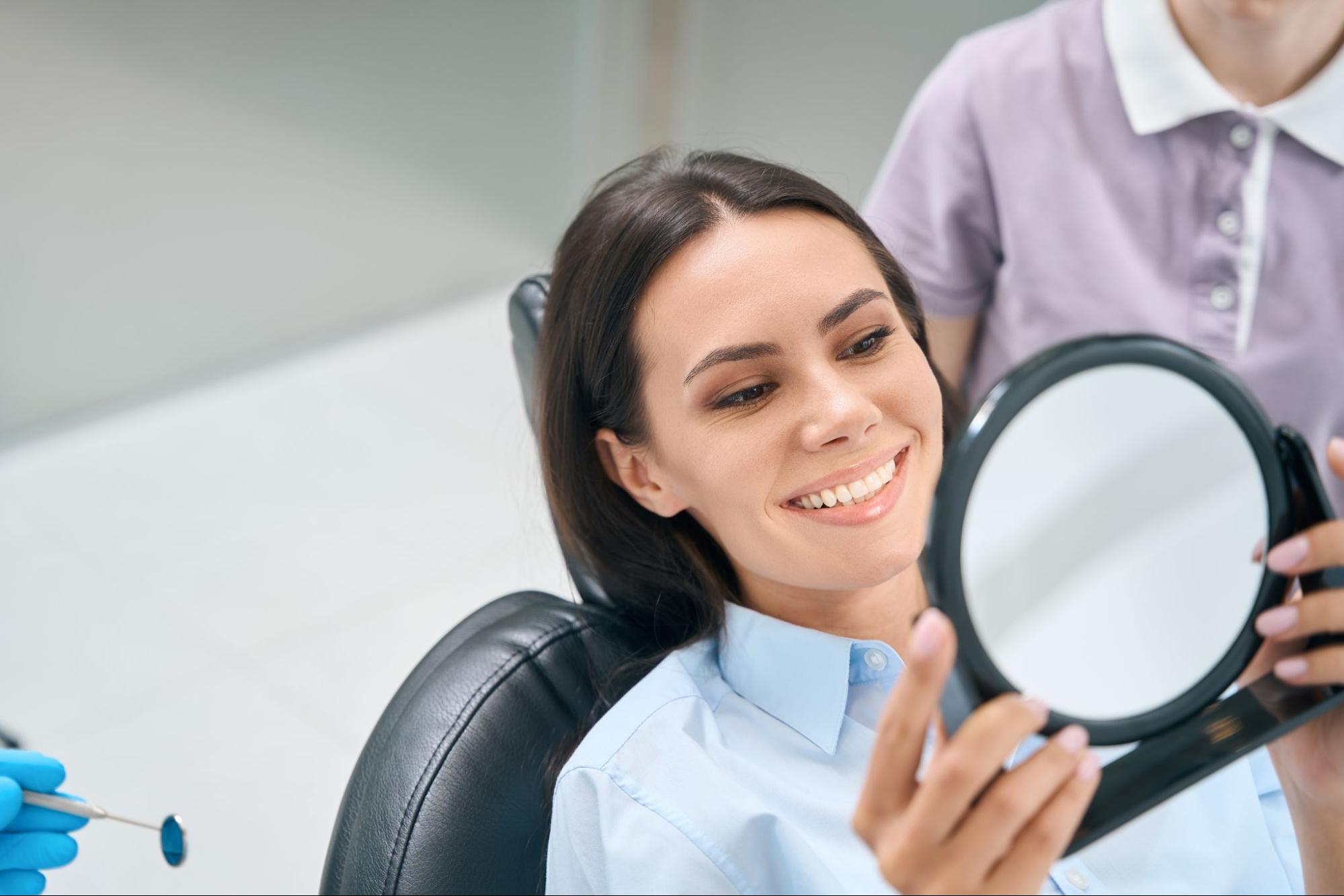 Woman in an exam chair smiling and looking at her teeth in a mirror. 