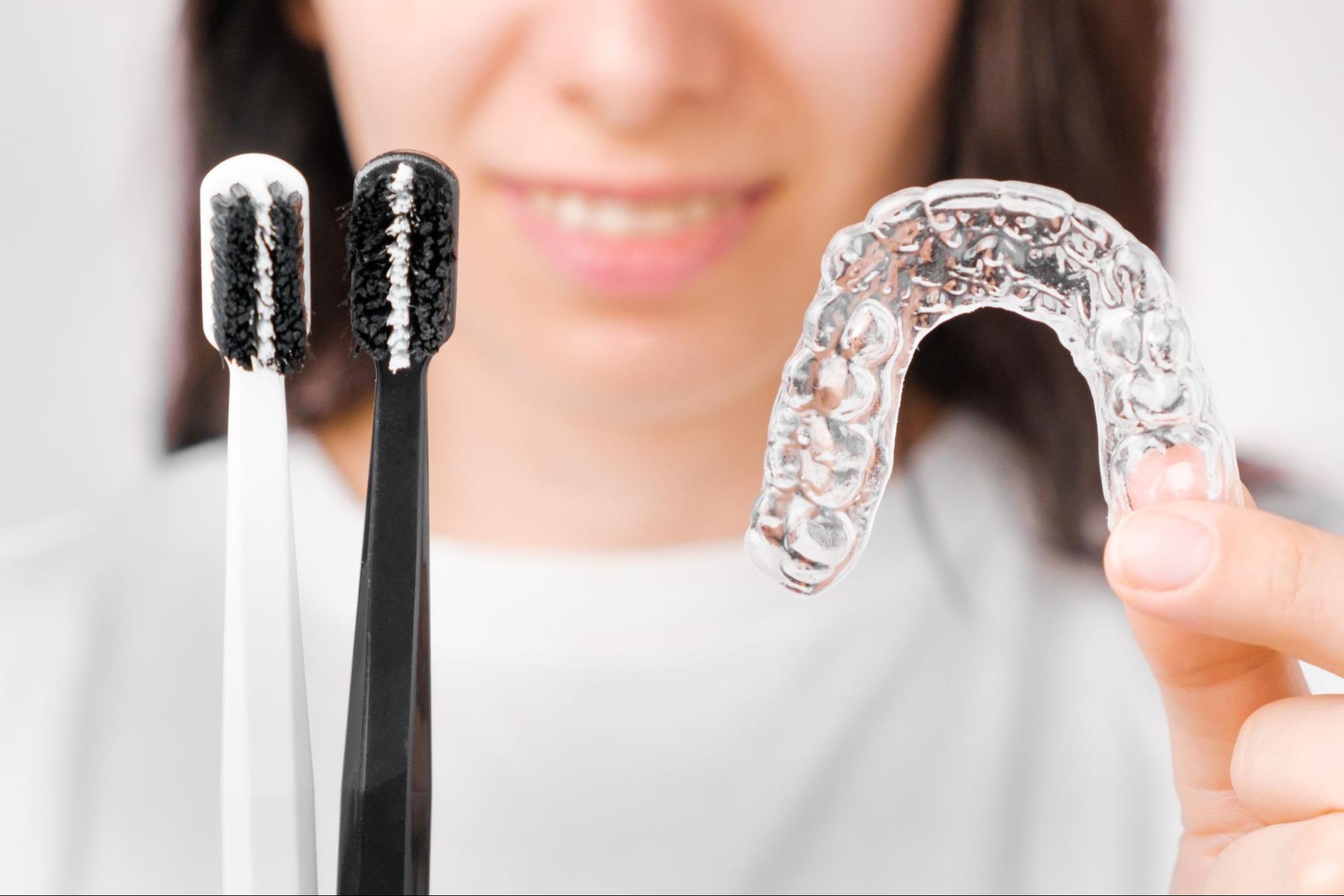 A woman holds black and white toothbrushes in her right hand and clear removable aligners in her left hand.