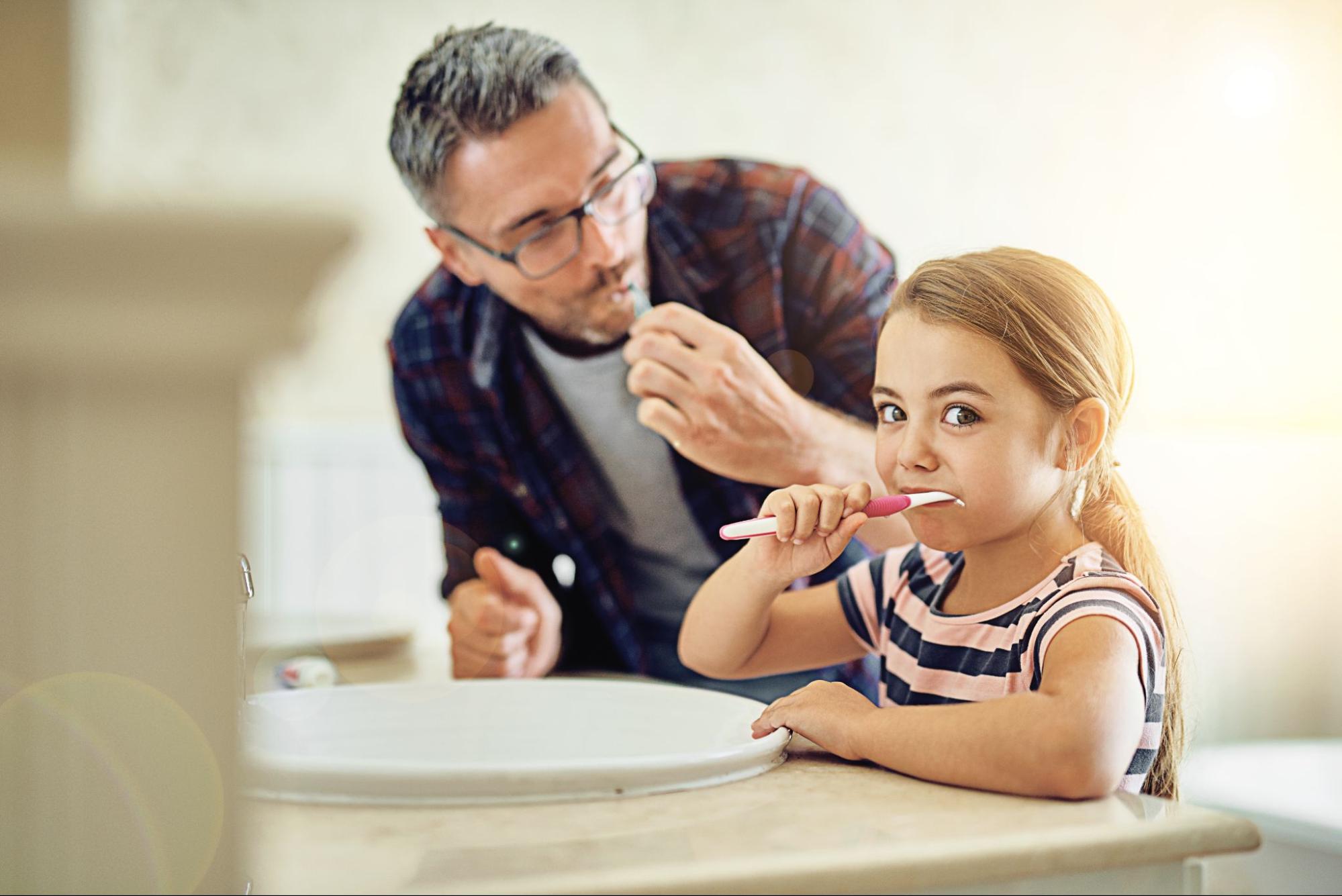 Young girl brushing her teeth with her dad at the sink.