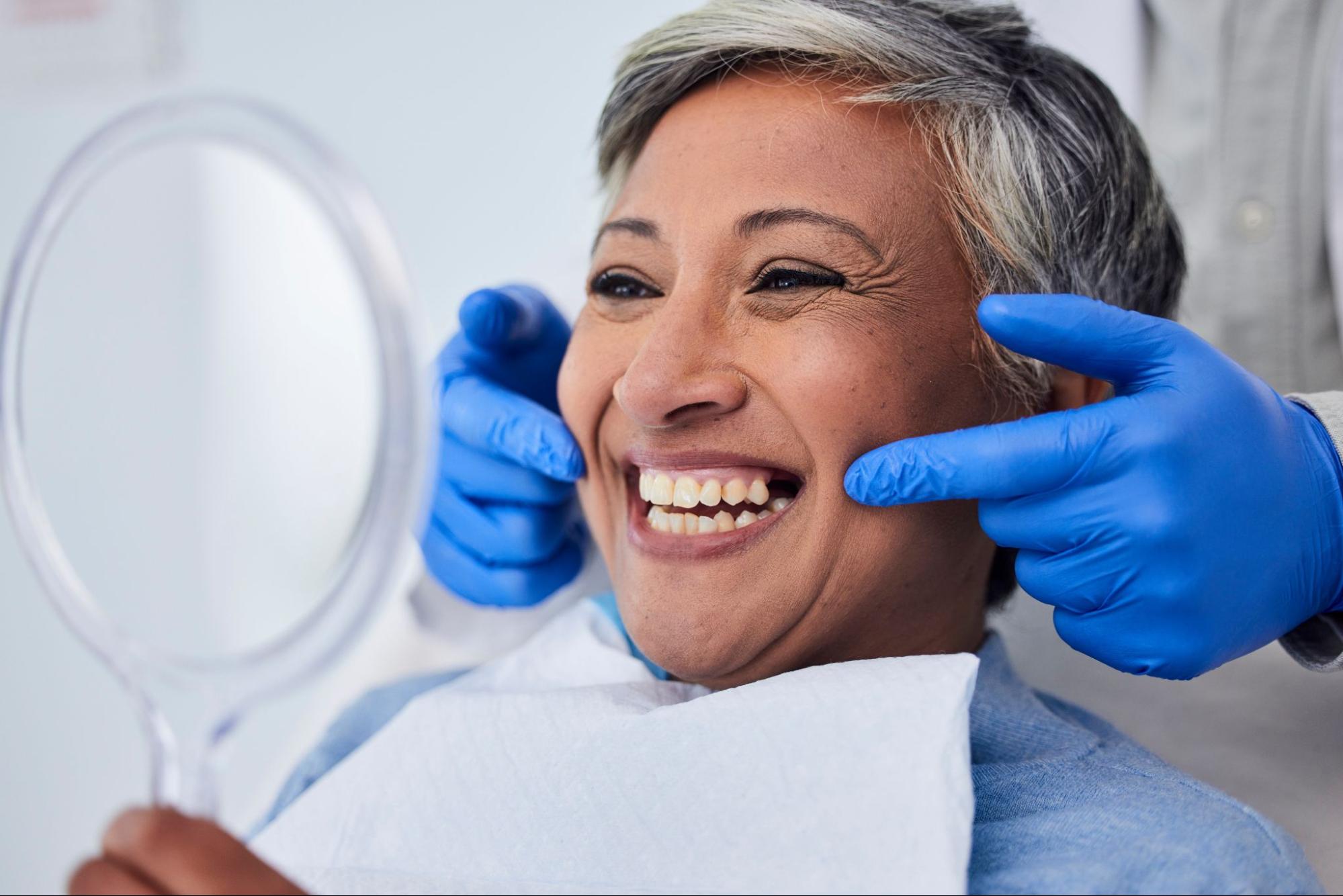 Senior woman receiving a dental consultation for implant recommendations, with the dentist’s gloved hands pointing to areas in her mouth needing restoration.