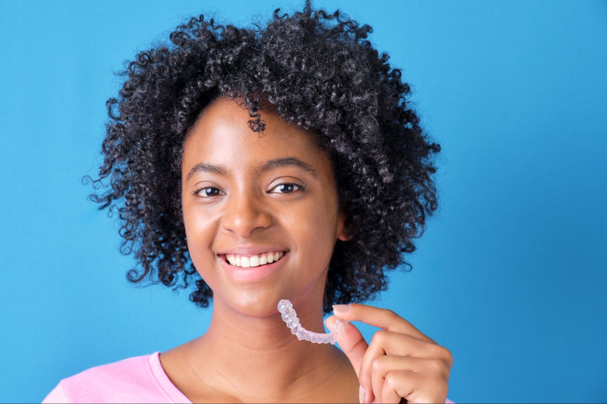 A Colombian woman with Afro hair smiles at the camera while holding an Invisalign® aligner.