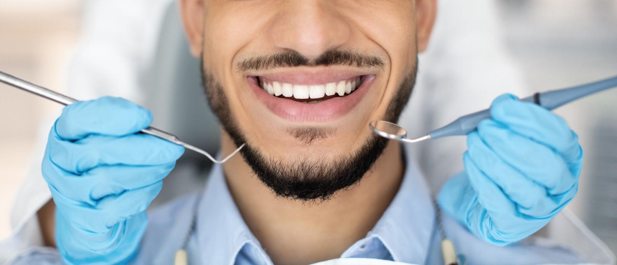 A close-up of a smiling, clean-shaven man showing his white teeth after a deep teeth cleaning procedure, with dental tools held near his mouth by unrecognized hands.