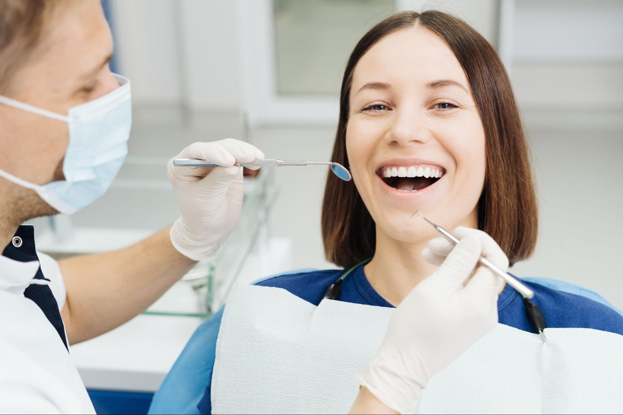 After a professional teeth whitening treatment, a dentist examines a happy female patient's mouth with professional instruments at a clinic.