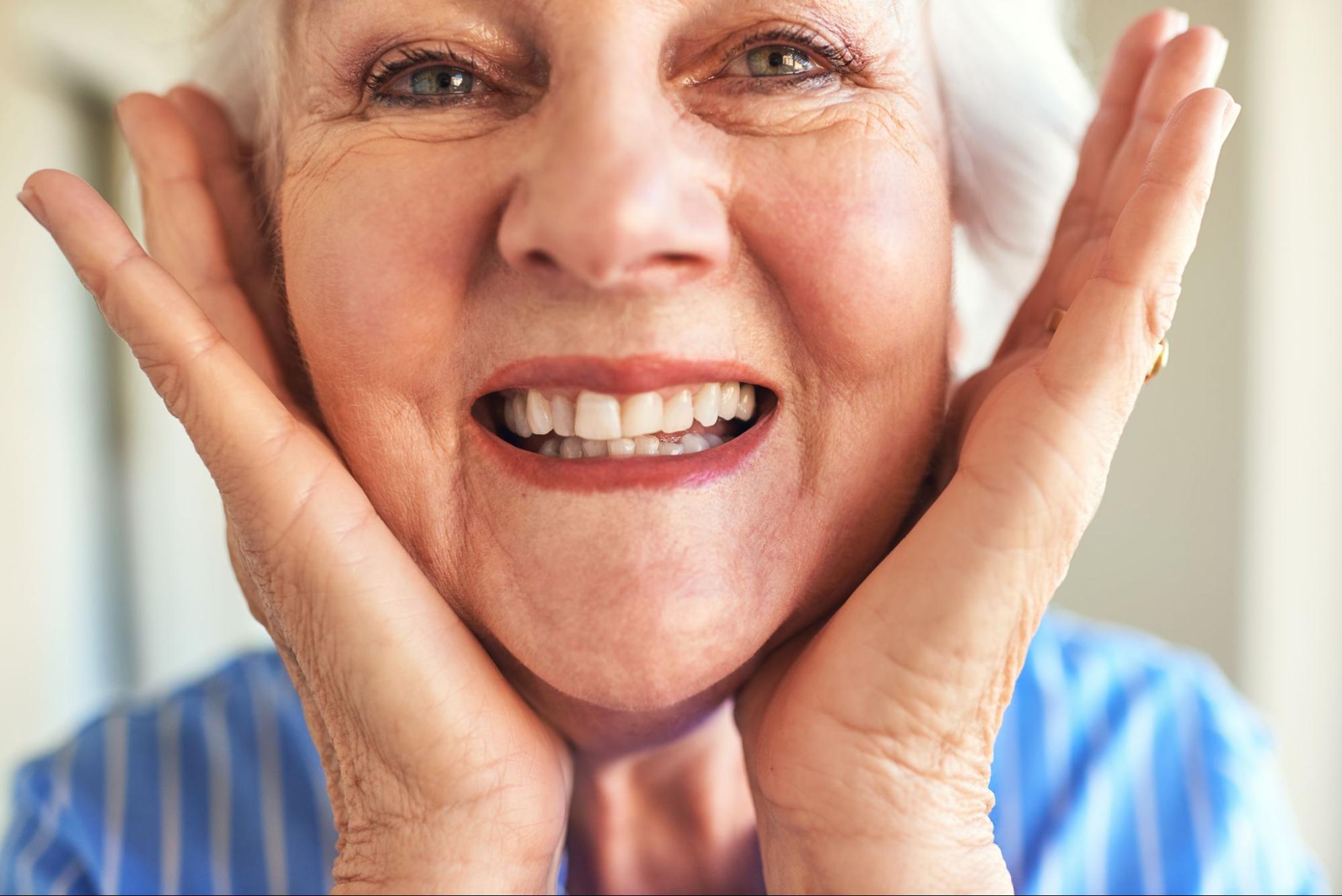 A close-up of a senior woman cupping her face and smiling proudly, showing off her new dental bridges.