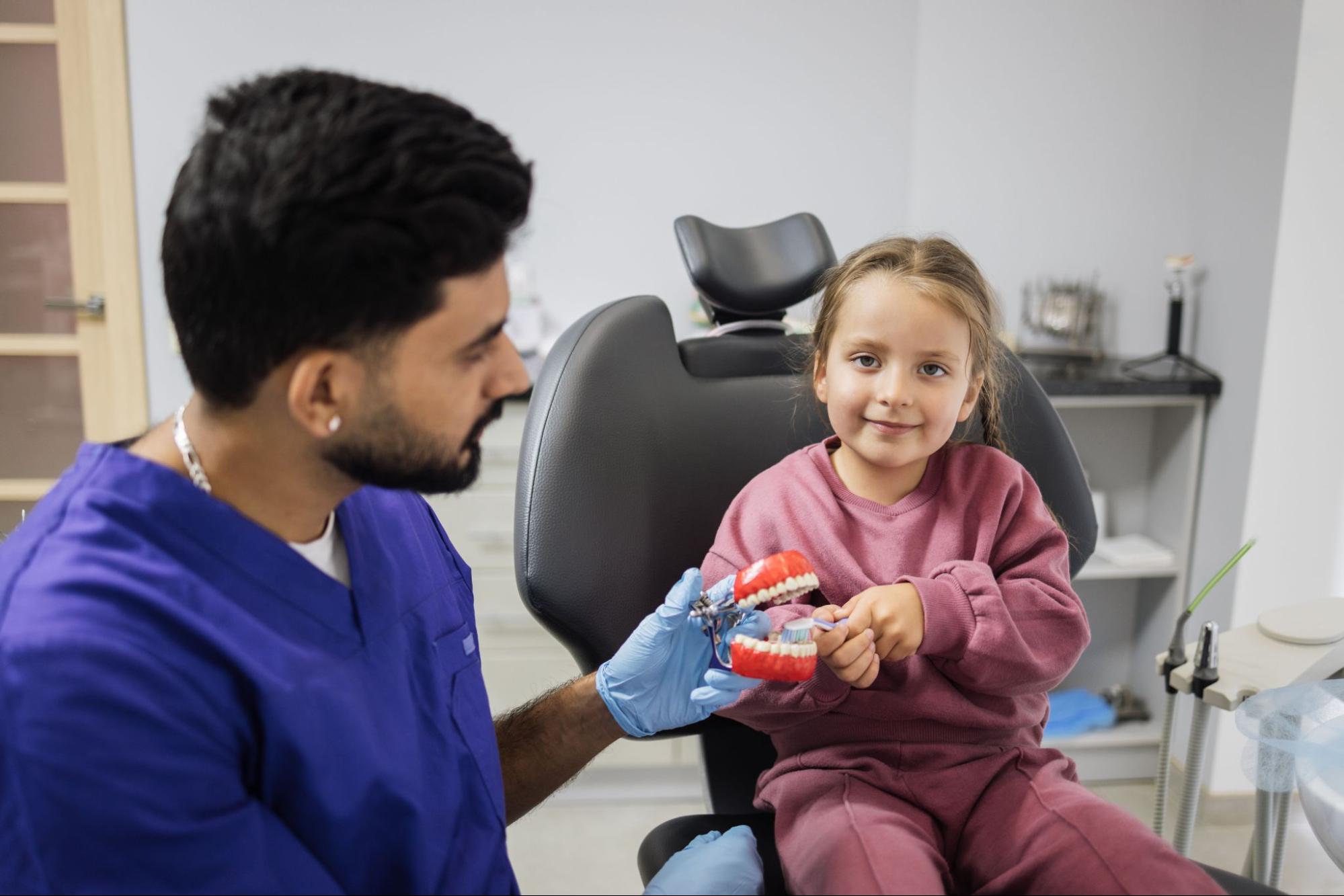 A male dentist for kids shows a smiling young girl how to brush her teeth using an artificial jaw model during a fun and educational dental visit.