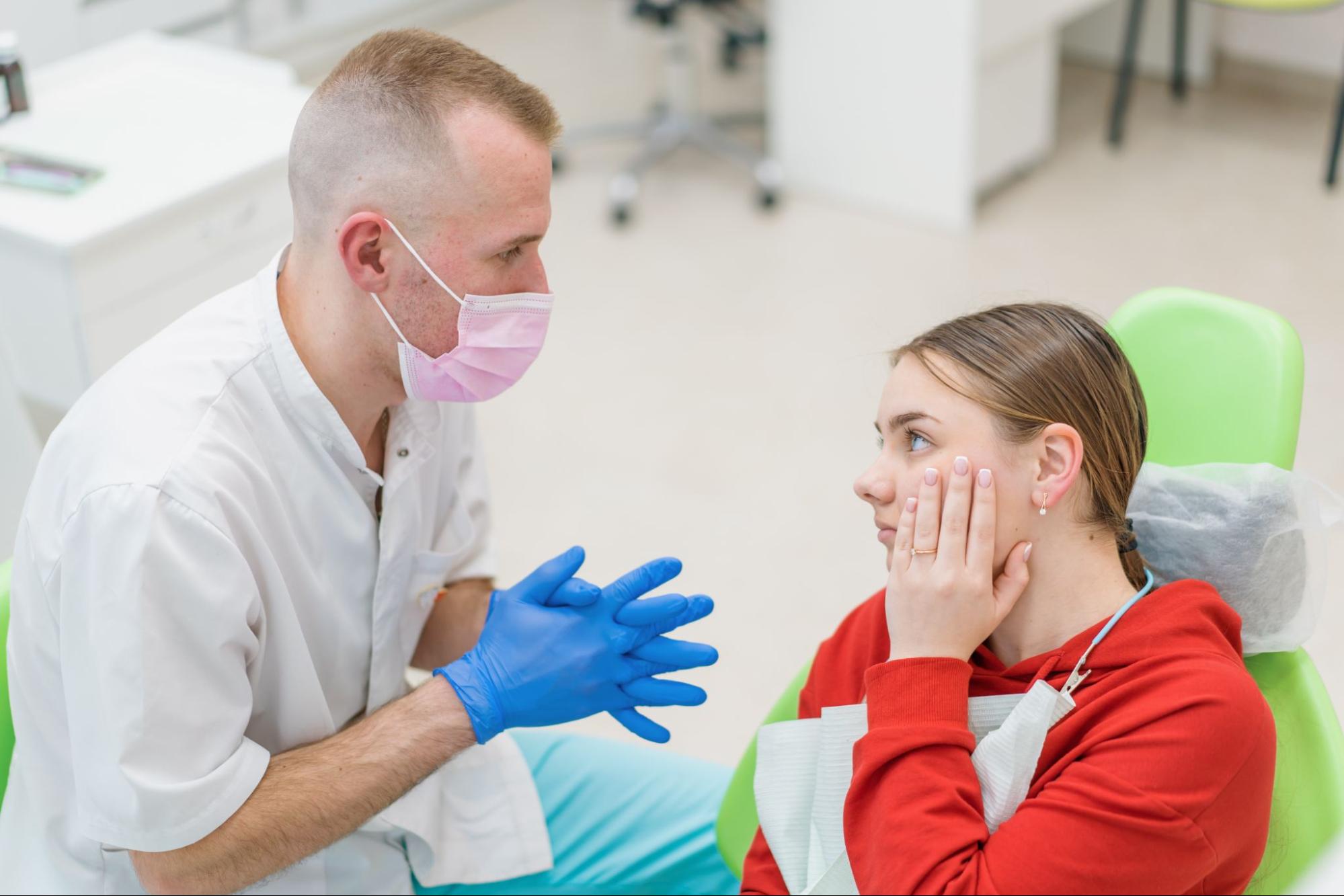 A doctor sitting beside a girl in an exam chair holding a hand to her face.
