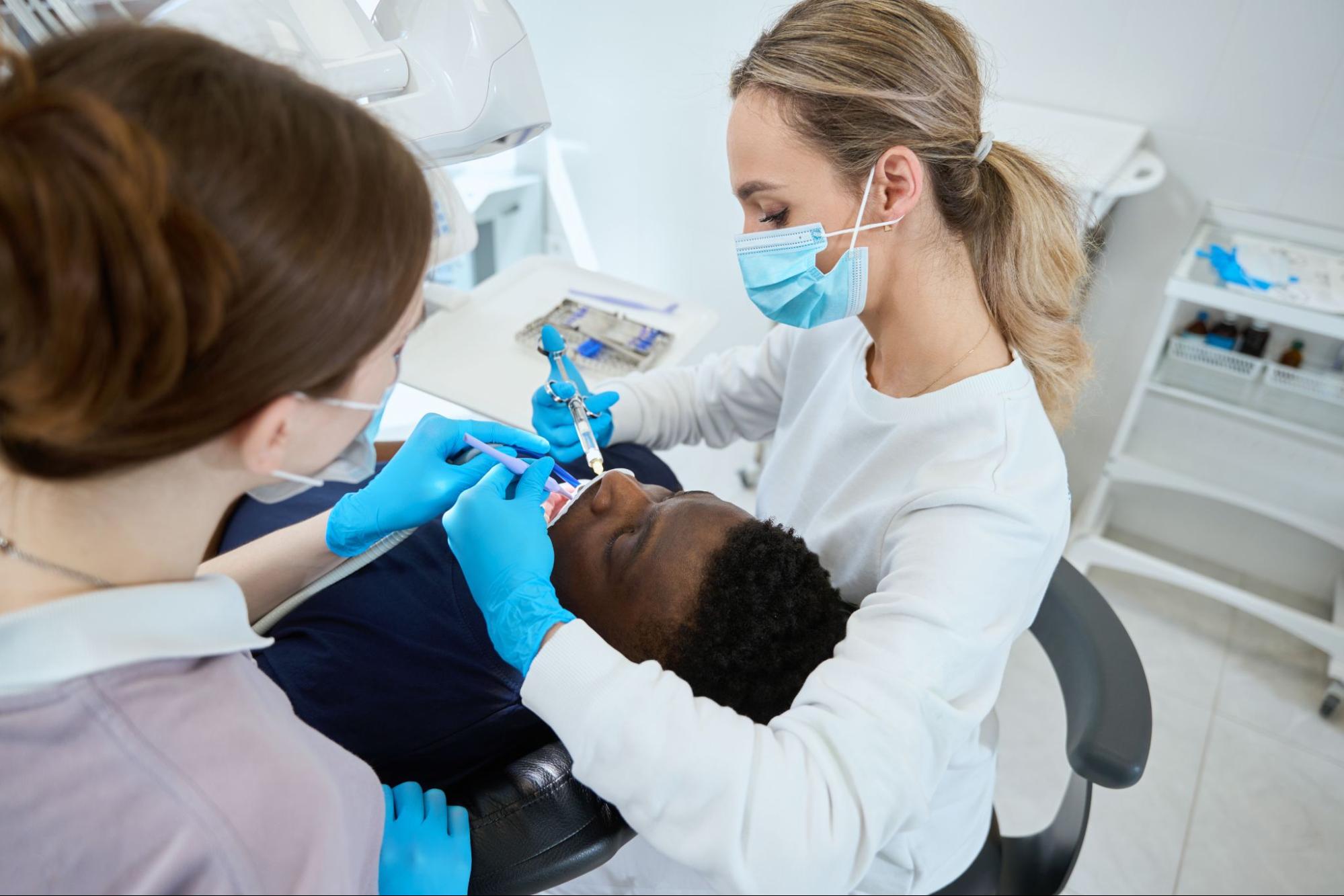 A female dentist thoroughly cleans the patient’s teeth before applying dental sealants, with a nurse assisting beside her.