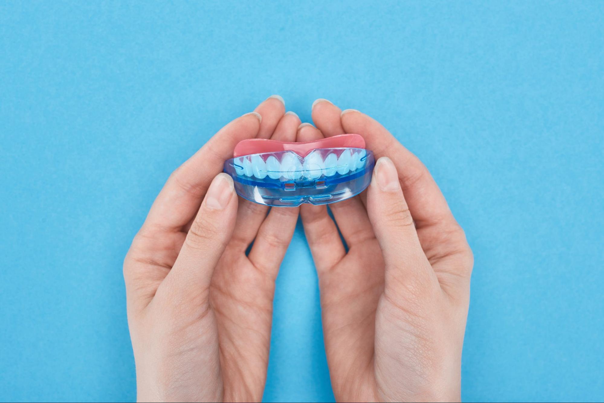 Hands holding a dental model with a clear blue night guard for teeth fitted on the upper arch.
