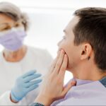 A man in a dentist office holding his cheek as he speaks to a female dental professional.