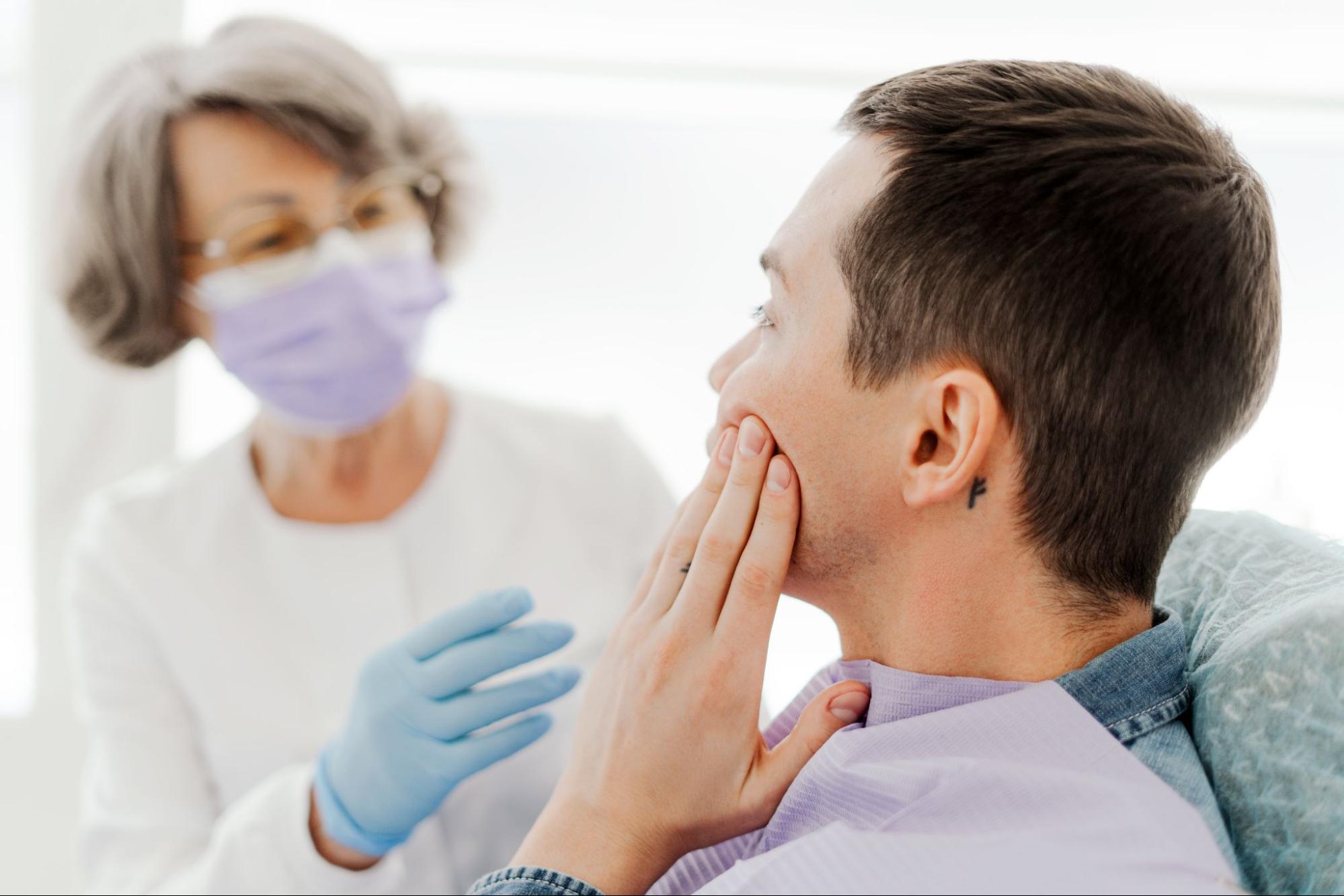 A man in a dentist office holding his cheek as he speaks to a female dental professional.