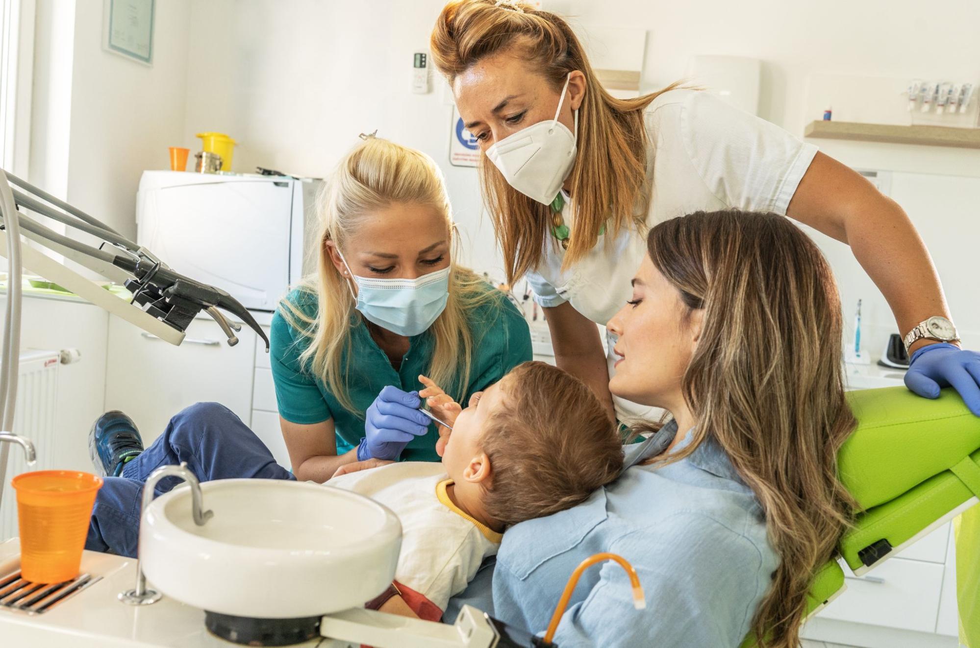 A pediatric dentist examines a young boy seated on his mother’s lap while a dental assistant looks on.