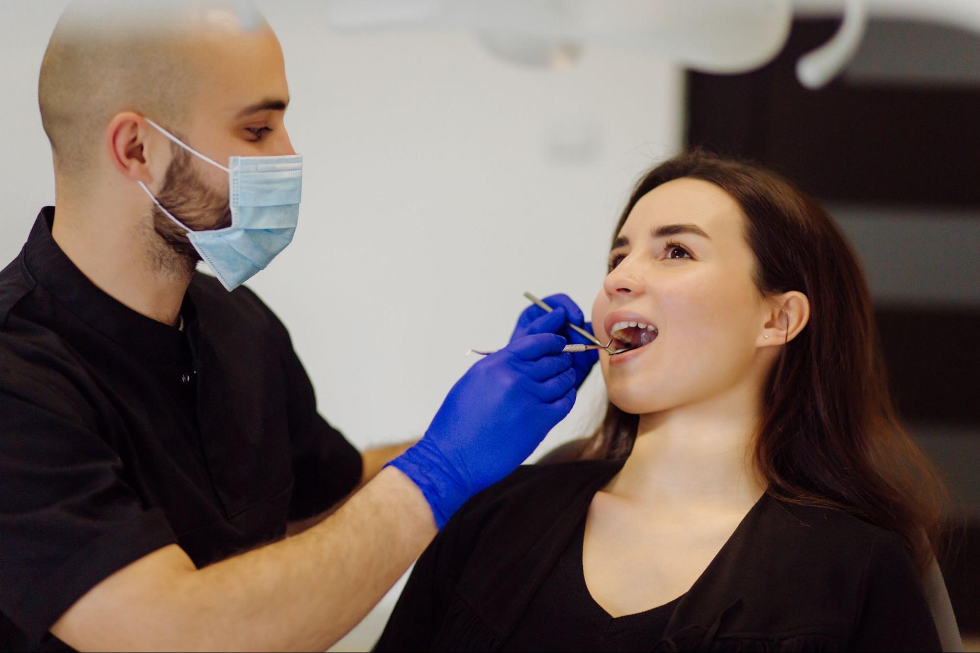 A masked male dentist closely examines a female patient’s gumline during a gum contouring evaluation.