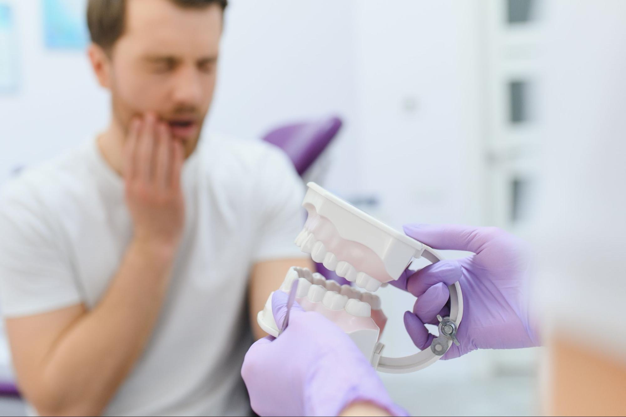Emergency dentist uses a jaw model to explain treatment to a man with visible tooth pain.