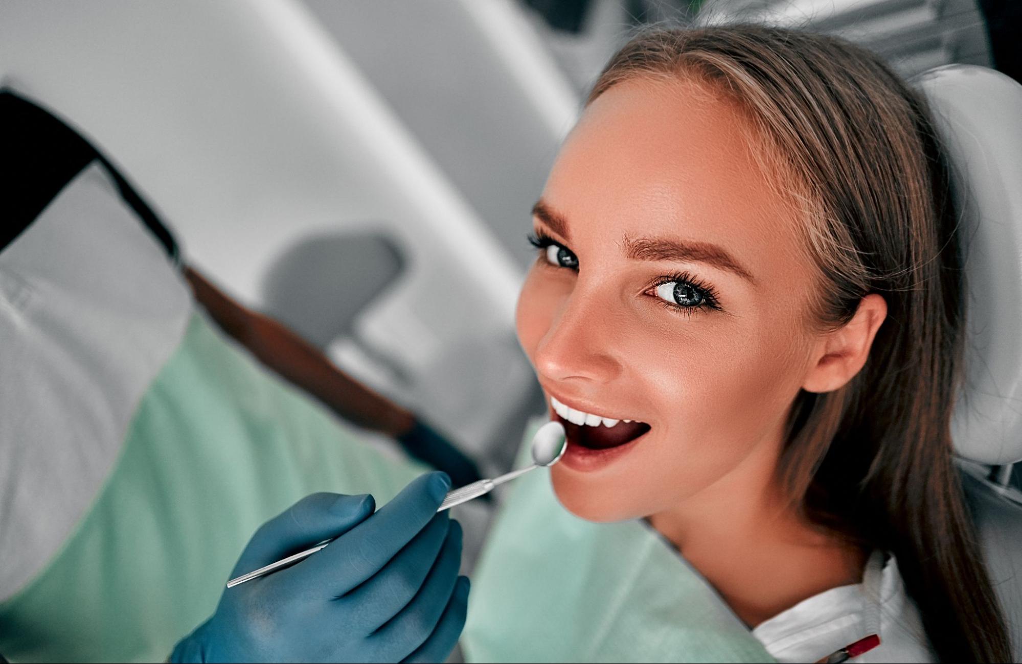 Woman smiling while lying down in a dental exam chair with a dental tool close to her mouth. 