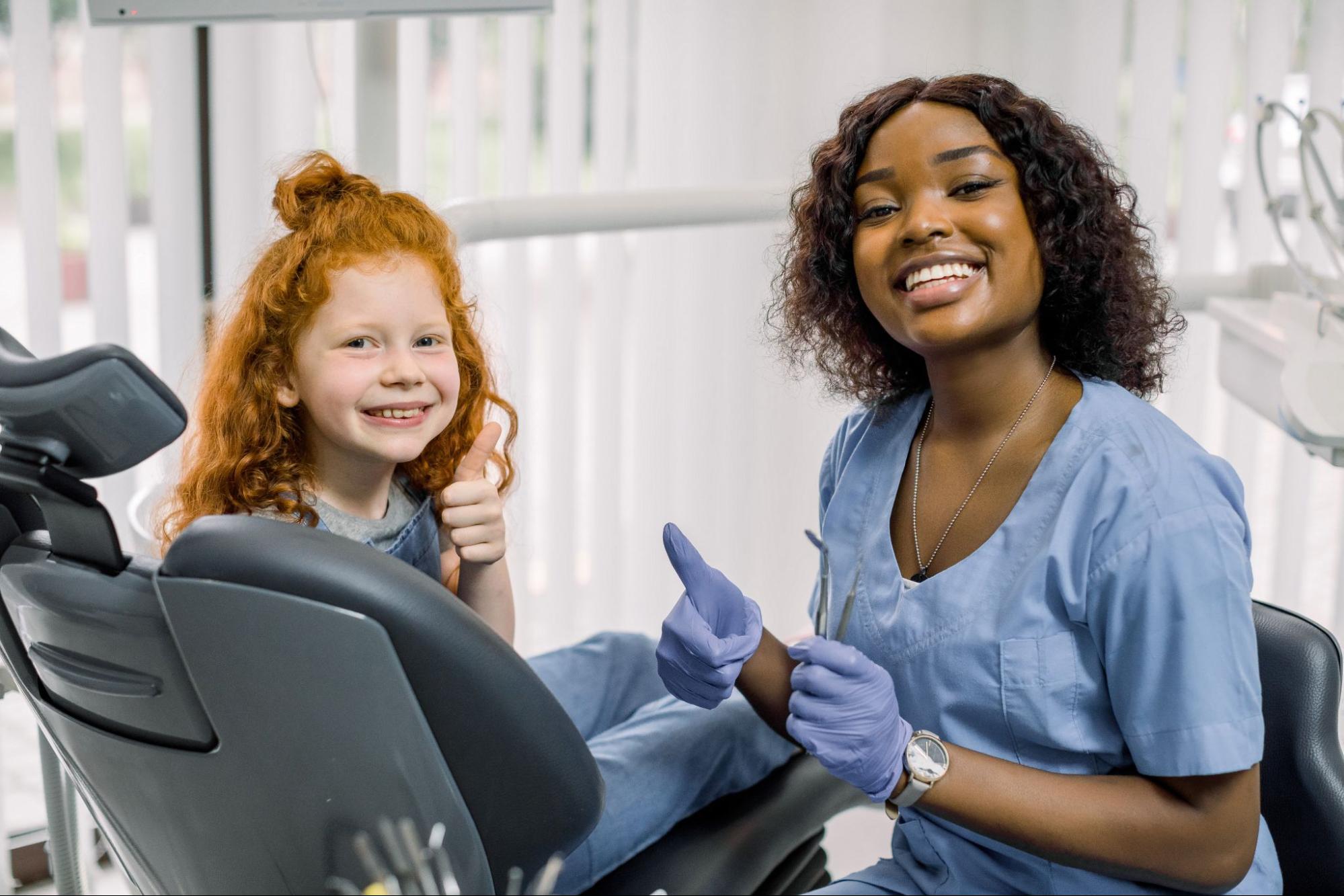 A cheerful young girl gives a thumbs up alongside her smiling female dentist, both expressing joy and confidence after a positive dental checkup.
