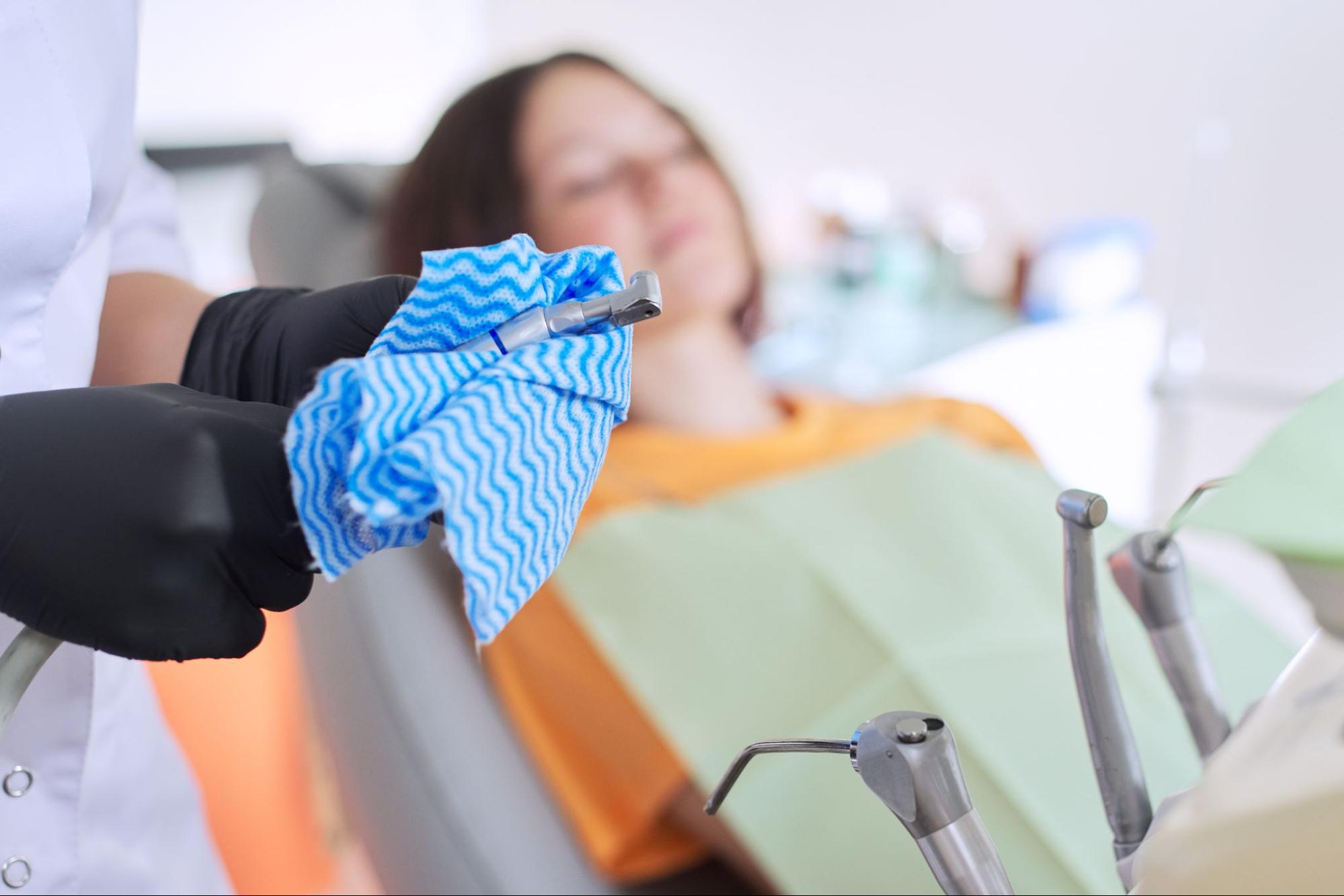 A close-up of a dental tool being prepared for teeth cleaning, with a blurred patient seated in the dental chair in the background.
